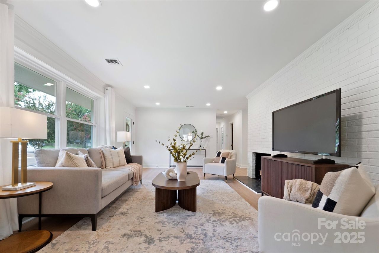 Bright living room with beige sofa, two white armchairs, large TV on wooden stand against white brick wall, oval wooden coffee table with vase, light rug, and large window with white curtains.
