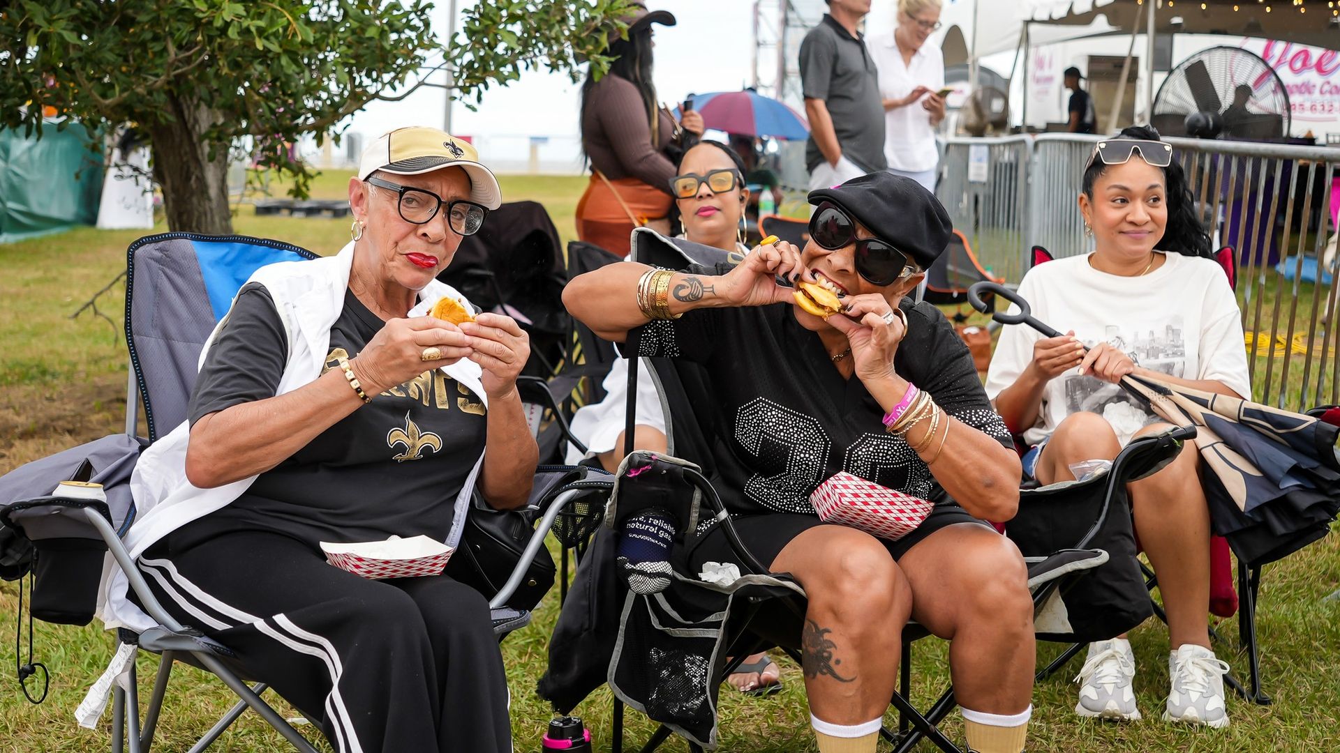 Group of women seated on folding chairs at an outdoor event, eating sandwiches. They wear sunglasses and hats; one in a tan cap, another in black. A white canopy tent and string lights are behind.