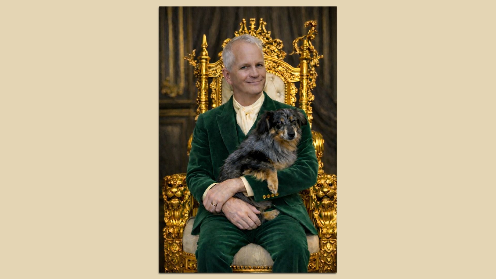 A man in green velvet suit sitting on an ornate gold throne, holding a small black and brown dog, both facing forward in a regal indoor setting. 