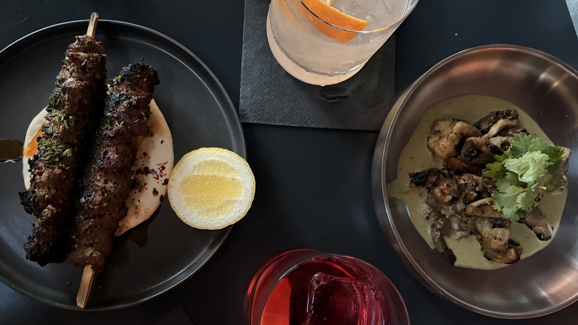 Top-down view of a dark table with two plates and two drinks: grilled meat skewers with lemon on the left; meat in green sauce with cilantro on the right; a red cocktail with an orange twist sits in front.