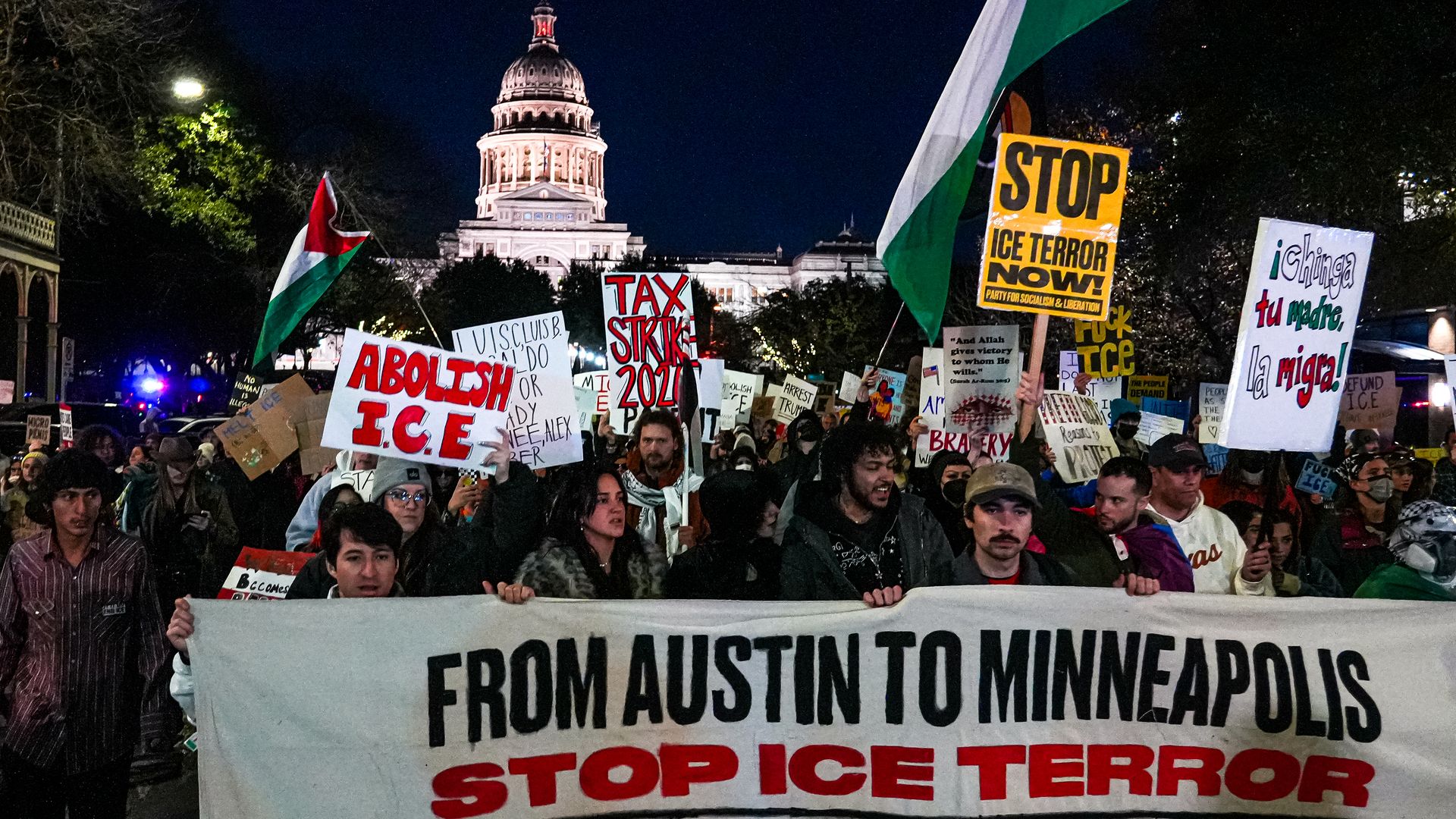 Nighttime protest in front of illuminated state capitol. Crowd holds signs including a large banner saying "From Austin to Minneapolis STOP ICE TERROR." Signs in red, yellow, white, green.