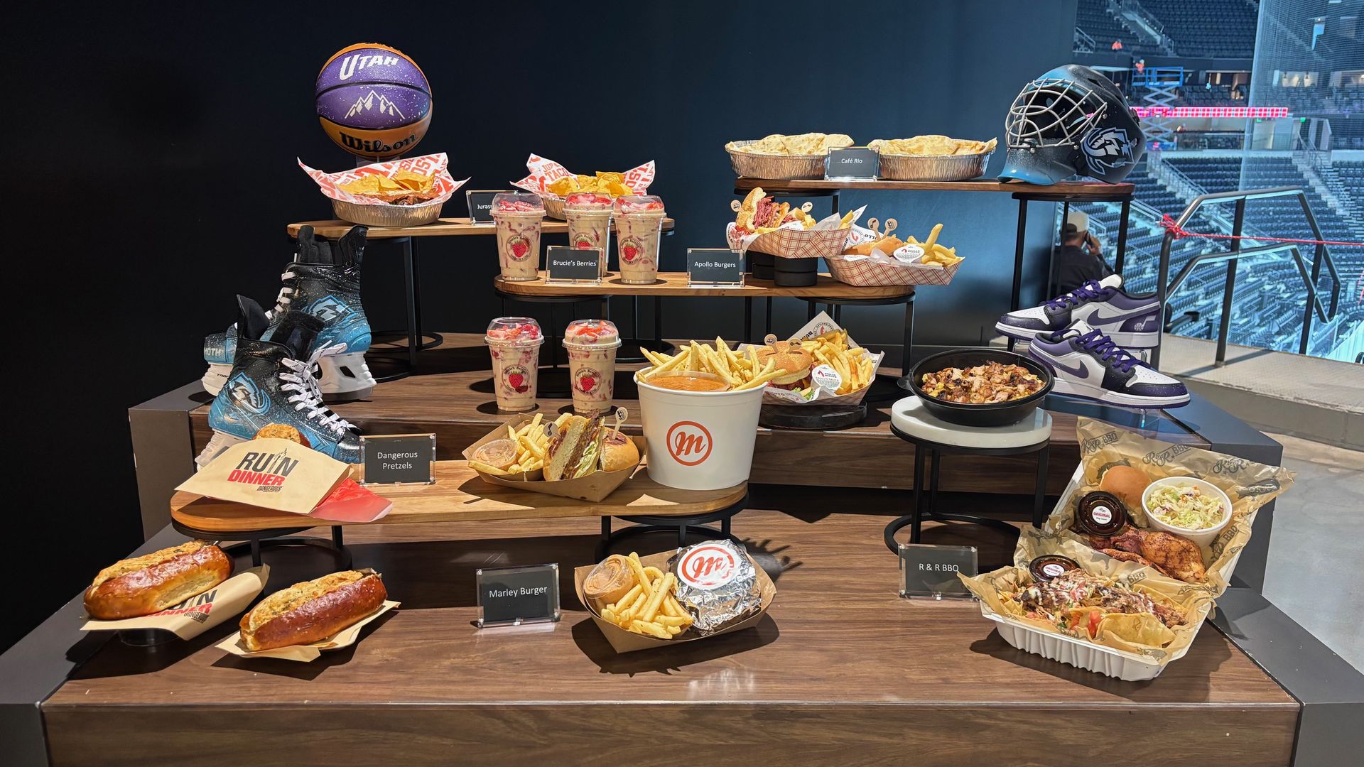 Sports-themed food display on wood tables featuring a purple Utah basketball, a hockey helmet and sneakers, with burgers, fries, nachos, pretzels, and drinks arranged in a stadium setting.