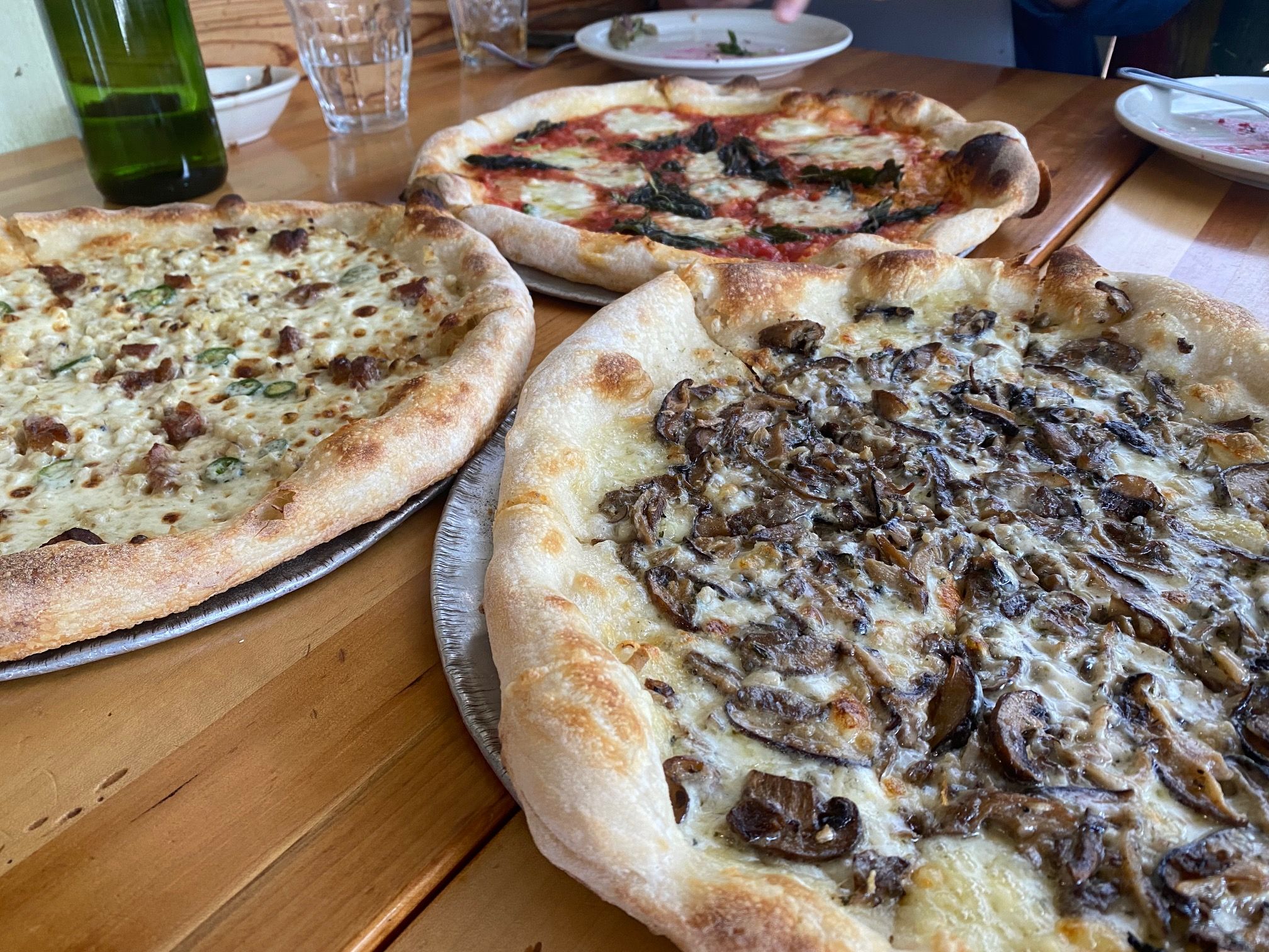 Three wood-fired pizzas on metal trays on a wooden table. One topped with mushrooms and cheese, another with white cheese and green peppers, and one with tomato, cheese, and basil.
