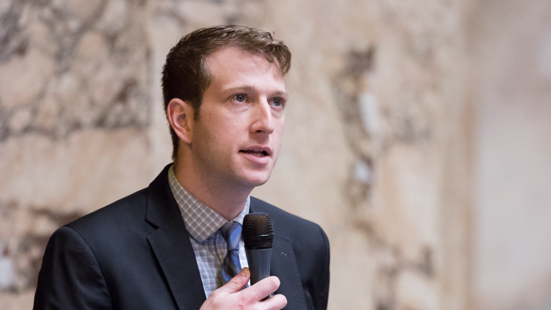 A man in a suit and tie speaks into a microphone in front of a marble wall.