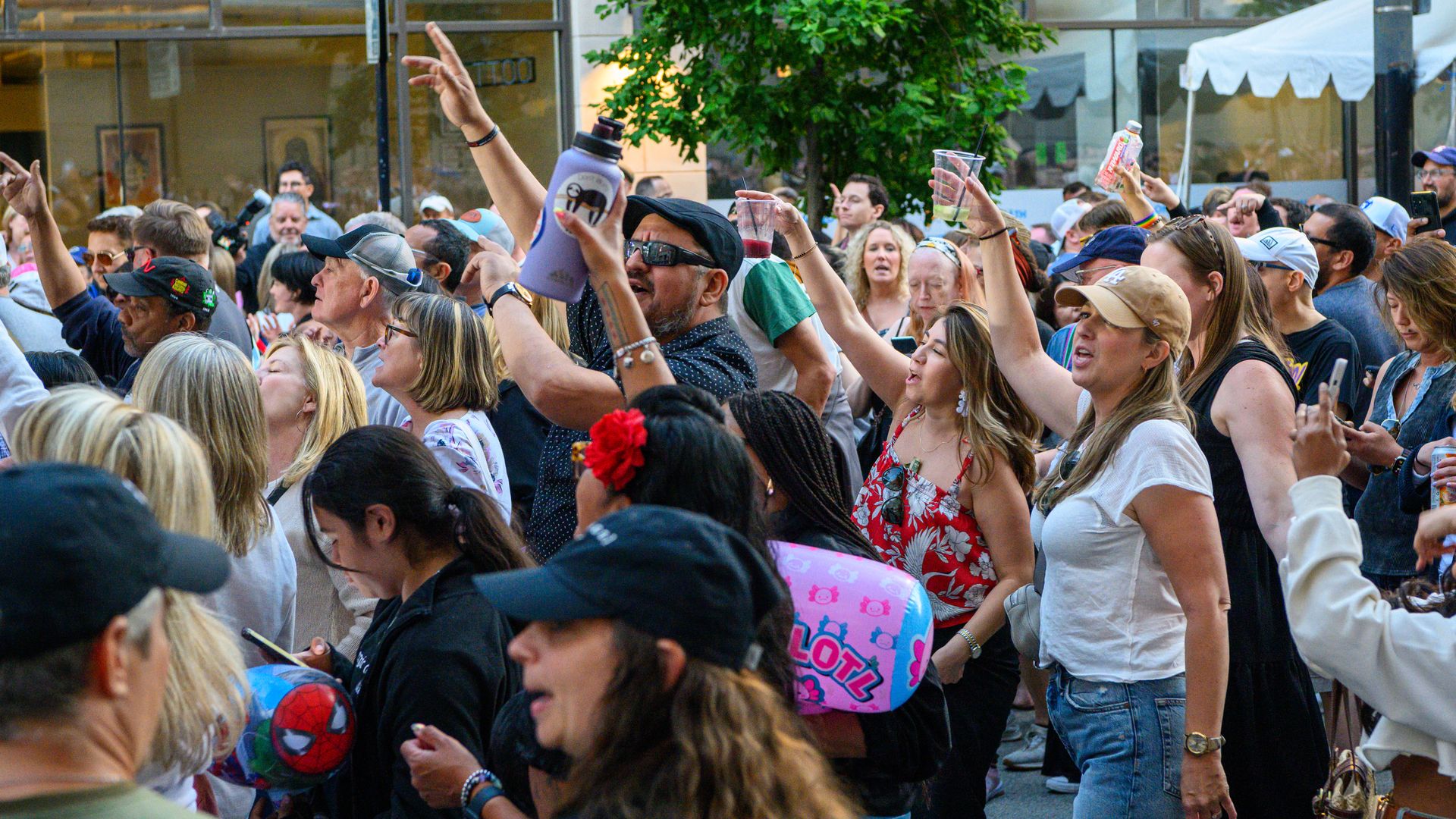 Crowded street festival with people raising cups and bottles amid a dense crowd. Many wear hats, sunglasses, and colorful clothing; glass storefronts and a leafy tree provide the backdrop.