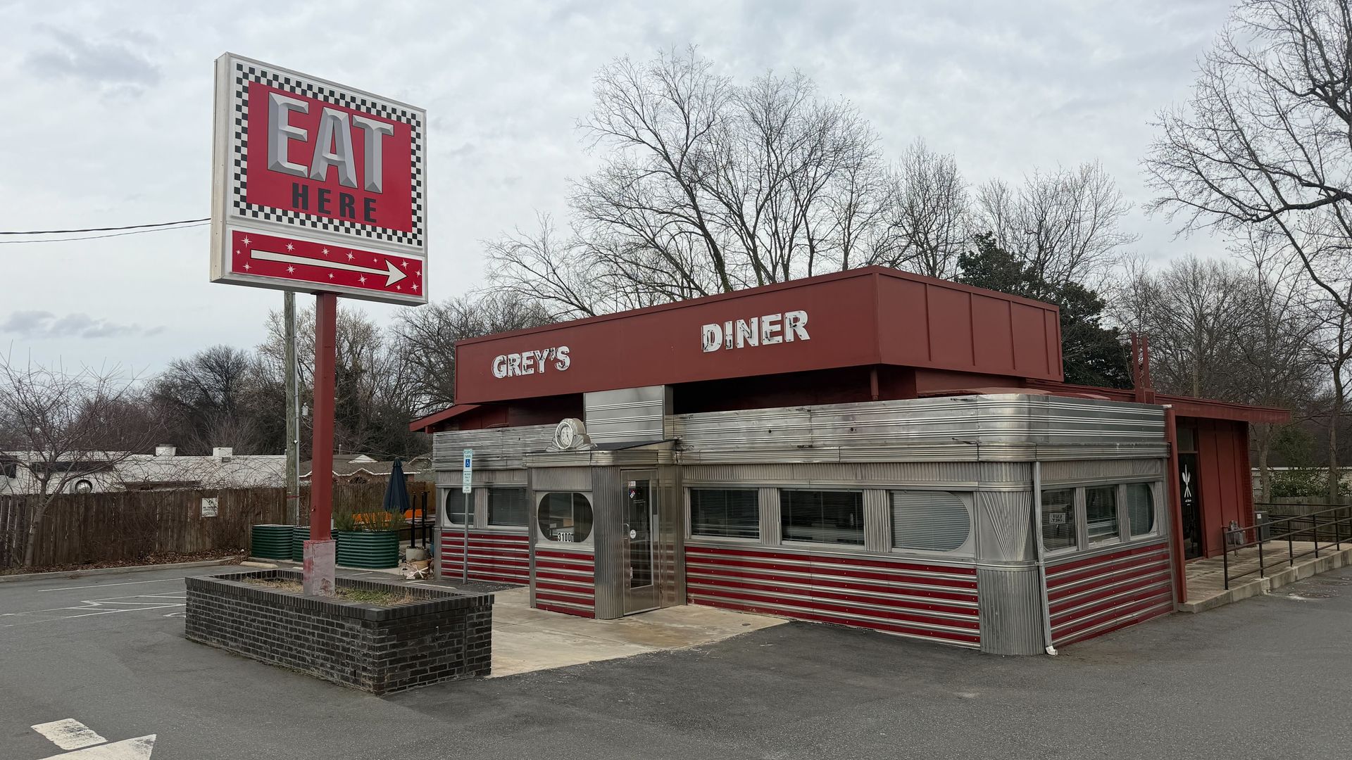 Grey's Diner with red and silver exterior under cloudy sky, large red sign saying EAT HERE with arrow pointing to diner, leafless trees in background.
