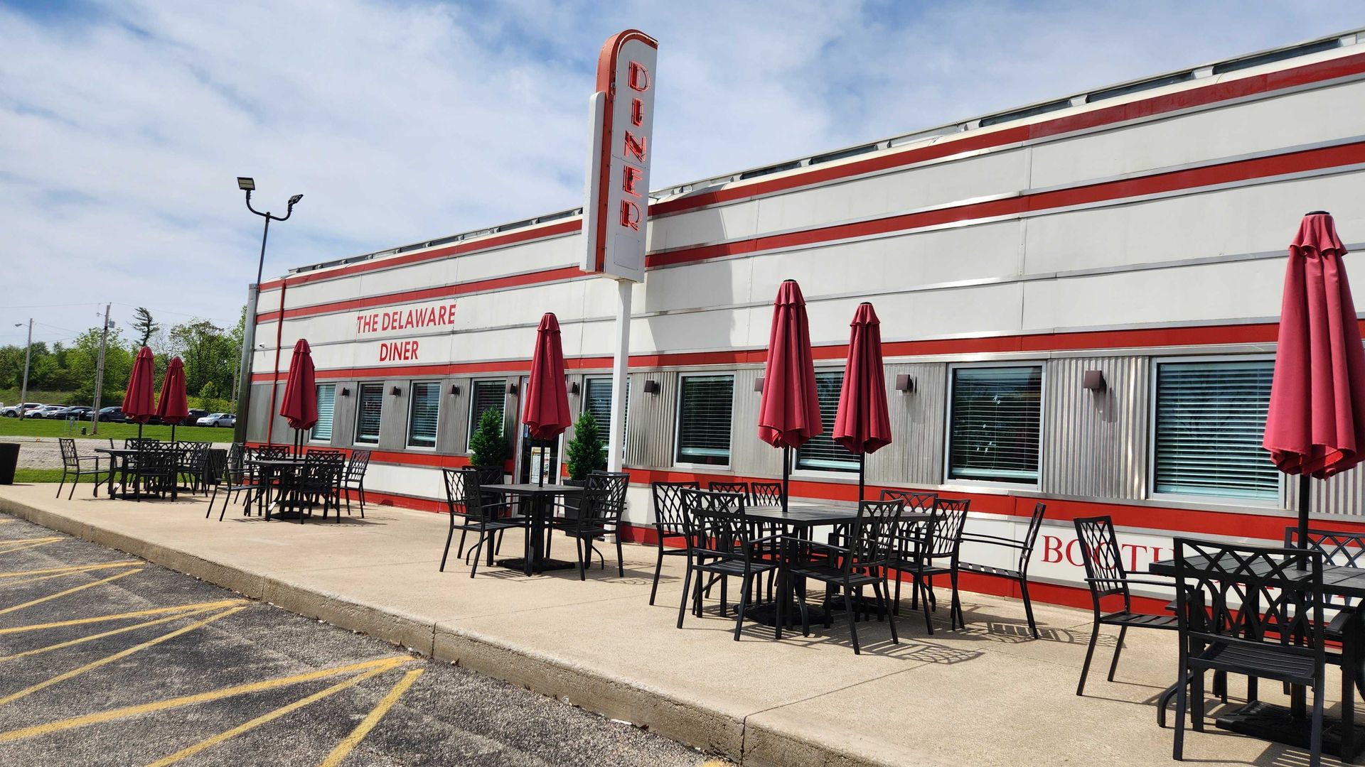 Exterior of The Delaware Diner: white building with red stripes, a vertical DINER sign, outdoor black metal tables with red umbrellas, and a blue sky with clouds.