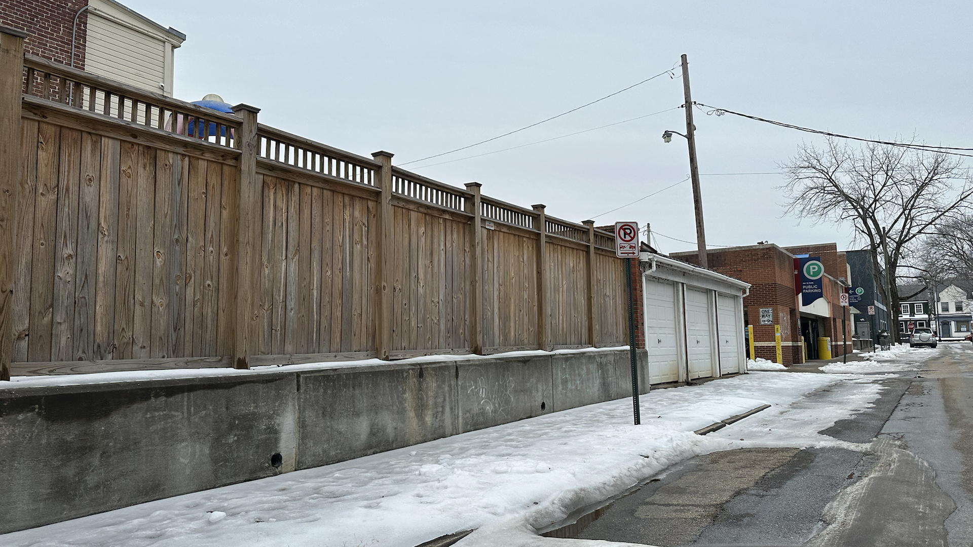 Snow-covered urban street with wooden fence, concrete base, utility pole, no parking sign, garages, public parking garage, bare tree, and overcast sky.