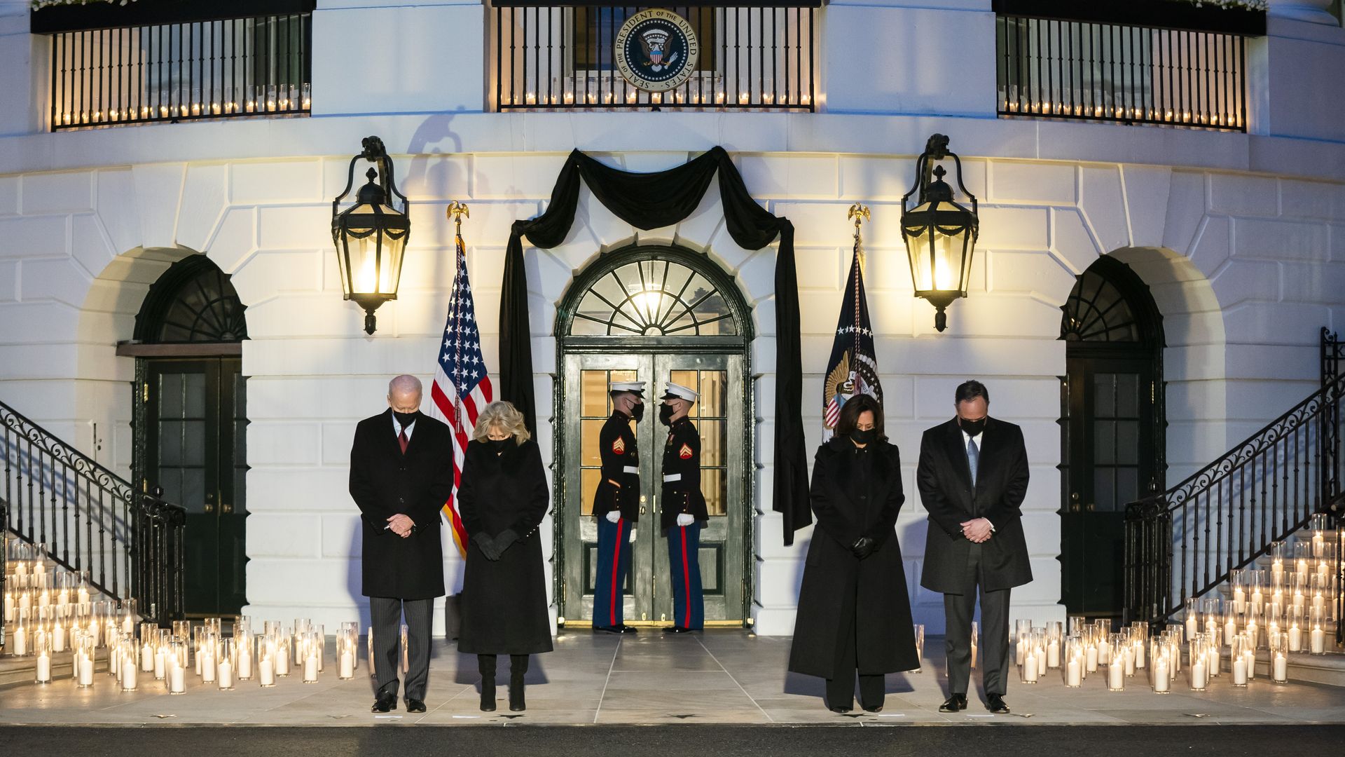 Photo of Joe Biden, Jill Biden, Kamala Harris and Doug Emhoff standing outside the White House with heads bowed and candles lit surrounding them
