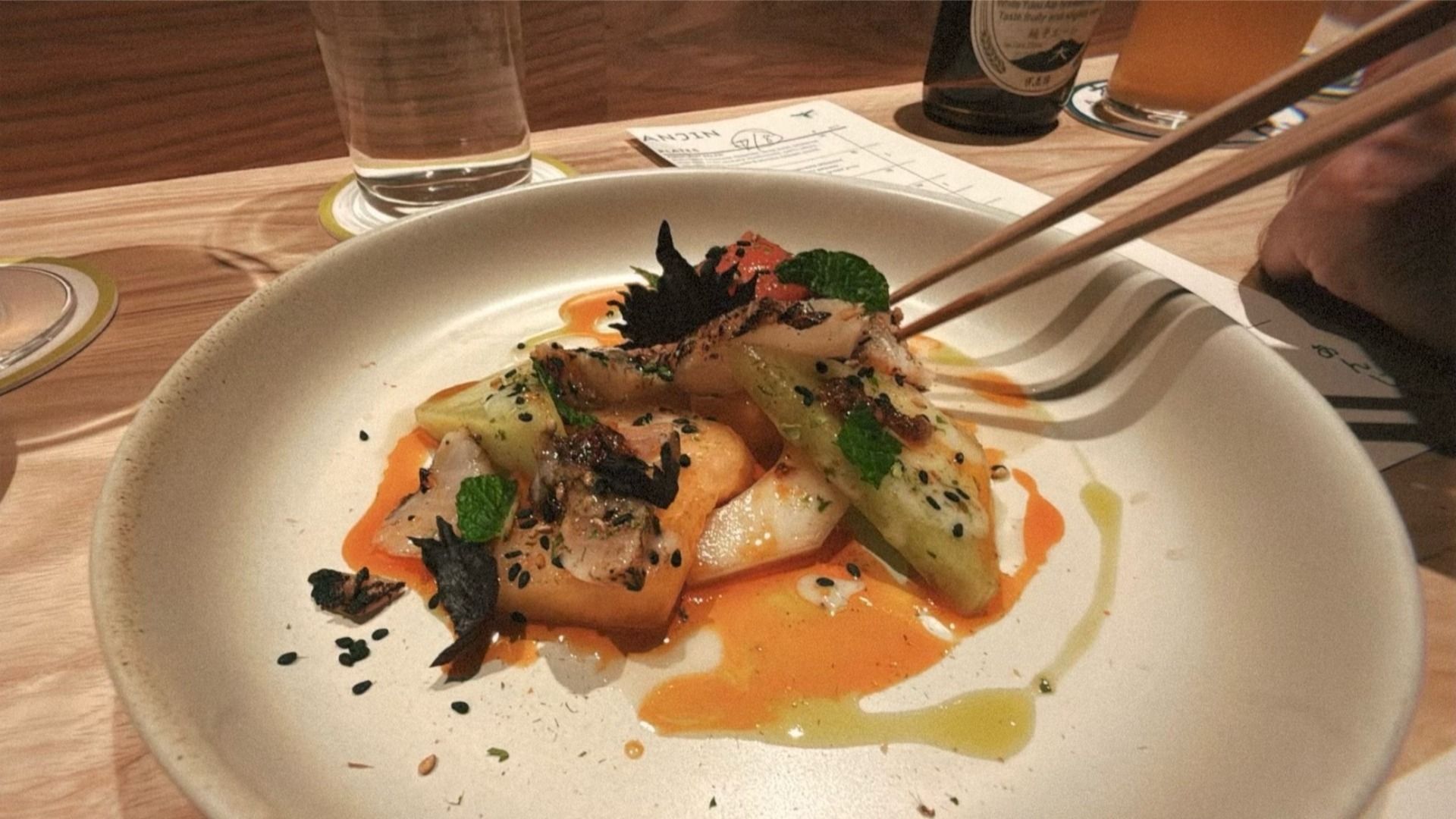 Plate with grilled vegetables and herbs garnished with black sesame seeds and drizzled with orange and green sauces, held by chopsticks on a wooden table at Anjin in Kansas City.