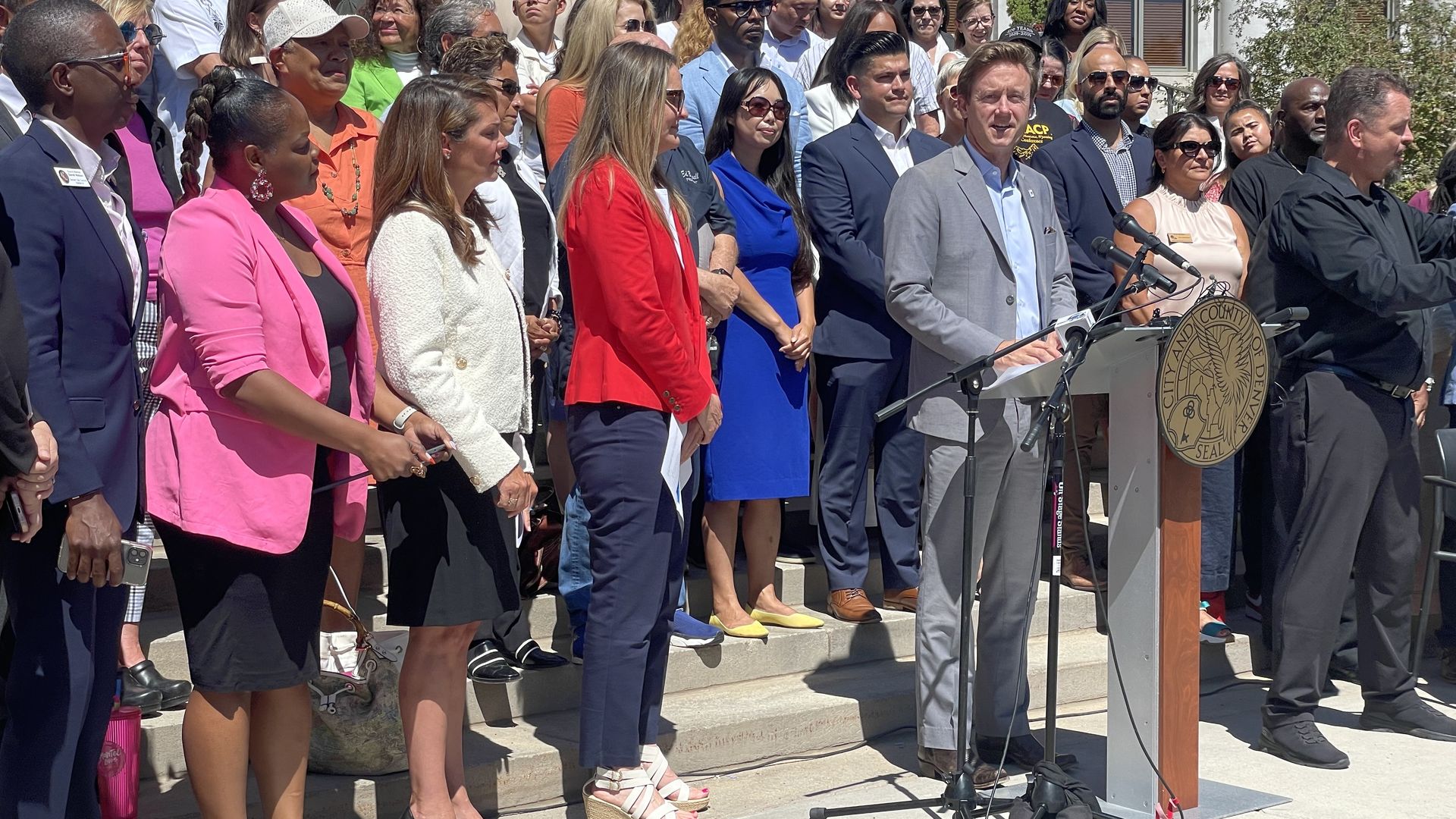 A man in a gray dress and a light blue shirt speaks at a lectern while surrounded by a large crowd. 