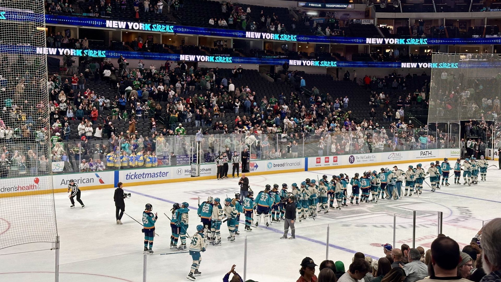 Professional Women's Hockey League players in teal and white uniforms line up on ice for post-game handshakes with cheering crowd in stands and electronic signs reading "NEW YORK SIRENS".