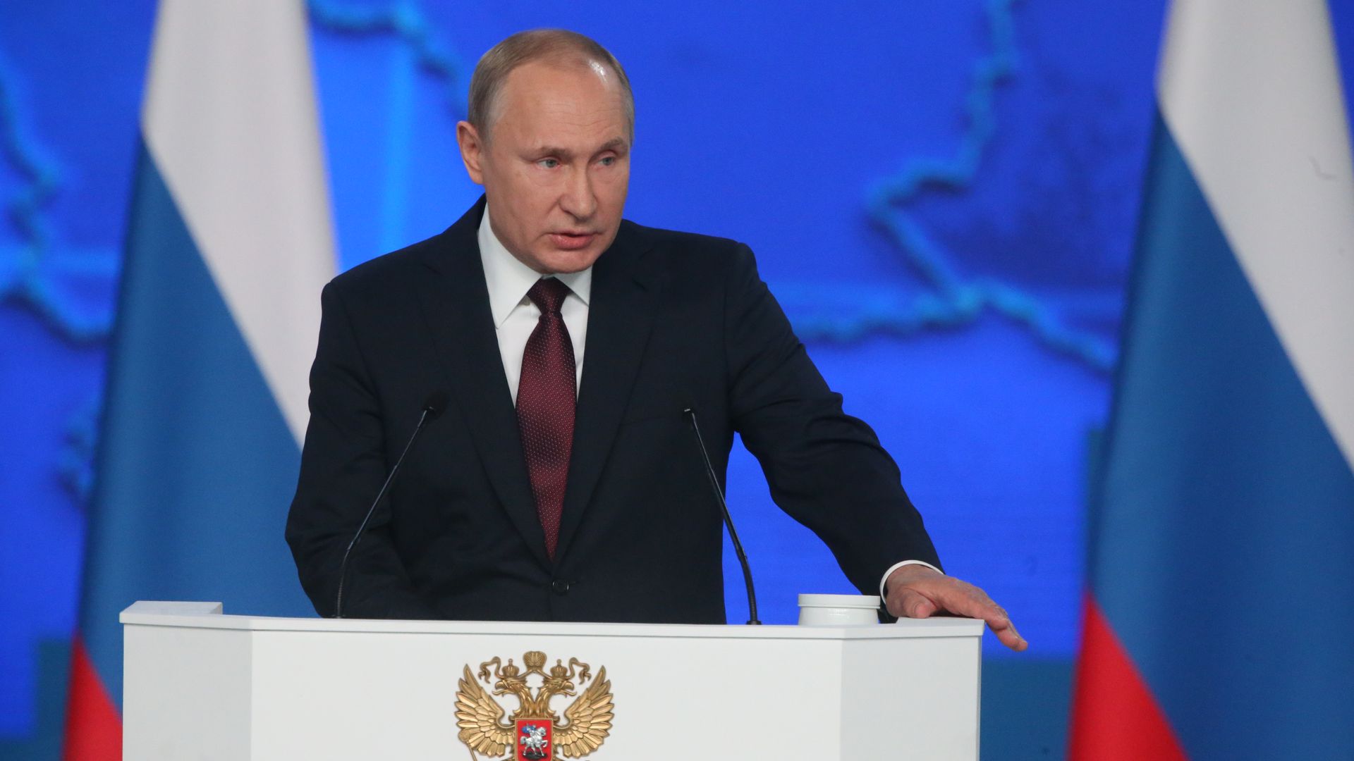In this image, Russian president Vladimir Putin stands in a suit and red tie at a white podium, in front of a bright blue background and a Russian flag.