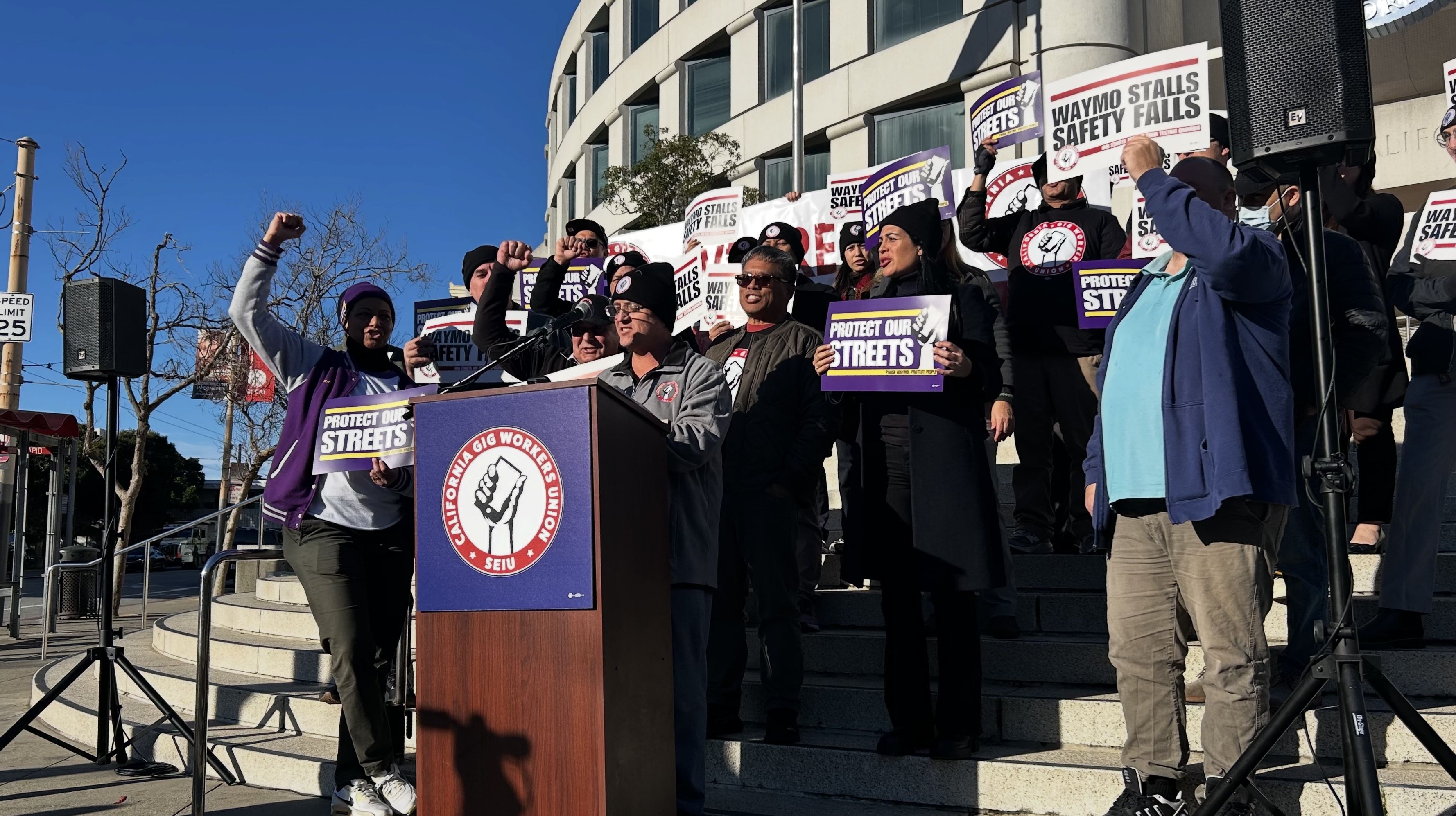 Group of diverse protesters with raised fists holding signs saying "Protect Our Streets" and "Waymo Stalls Safety Falls" at a California Gig Workers Union rally outside a building under clear blue sky.