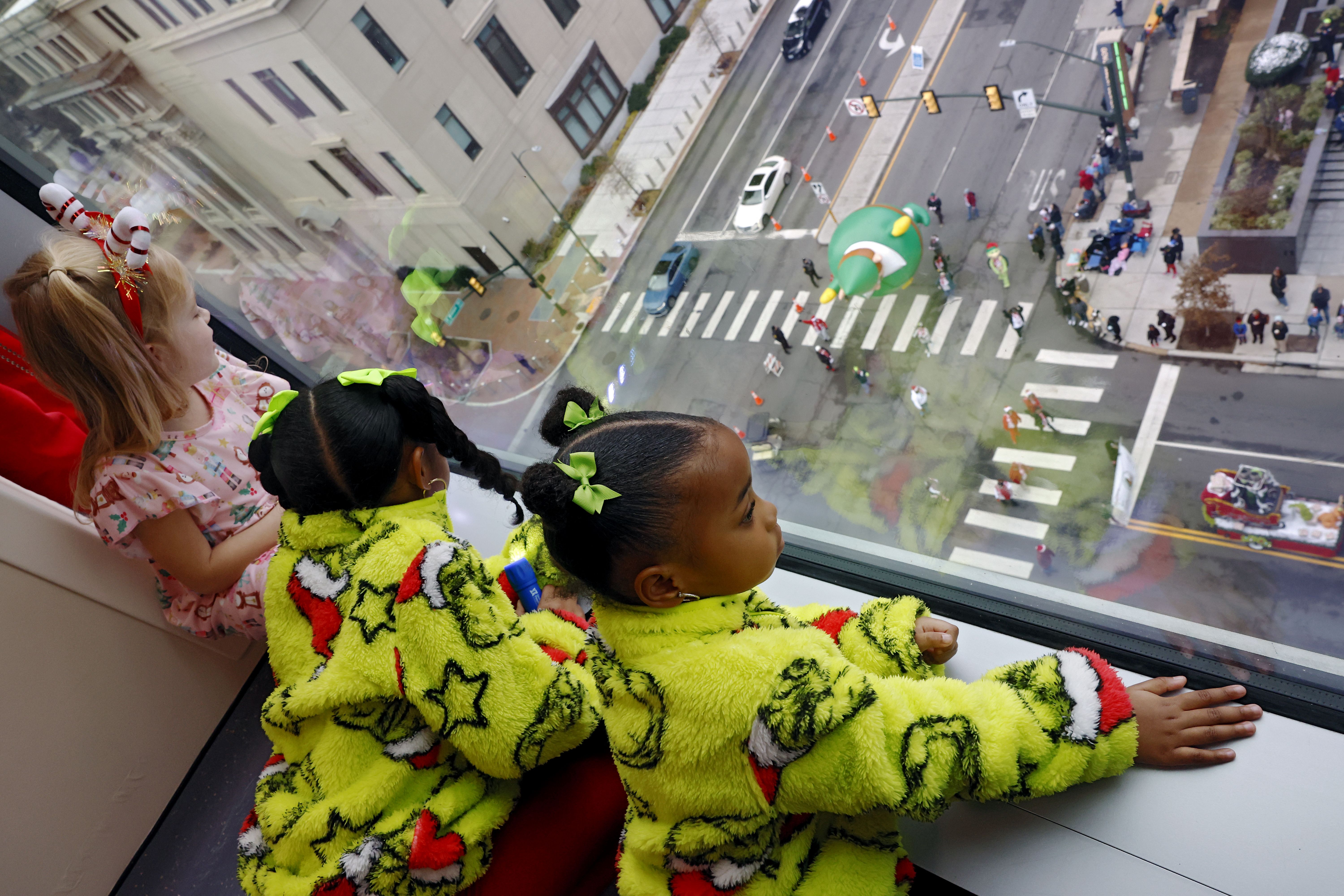 Three children in bright neon green fuzzy outfits peek from a window, watching a city street below with a big green dragon balloon, parked cars, pedestrians, and crosswalk lines during a parade.