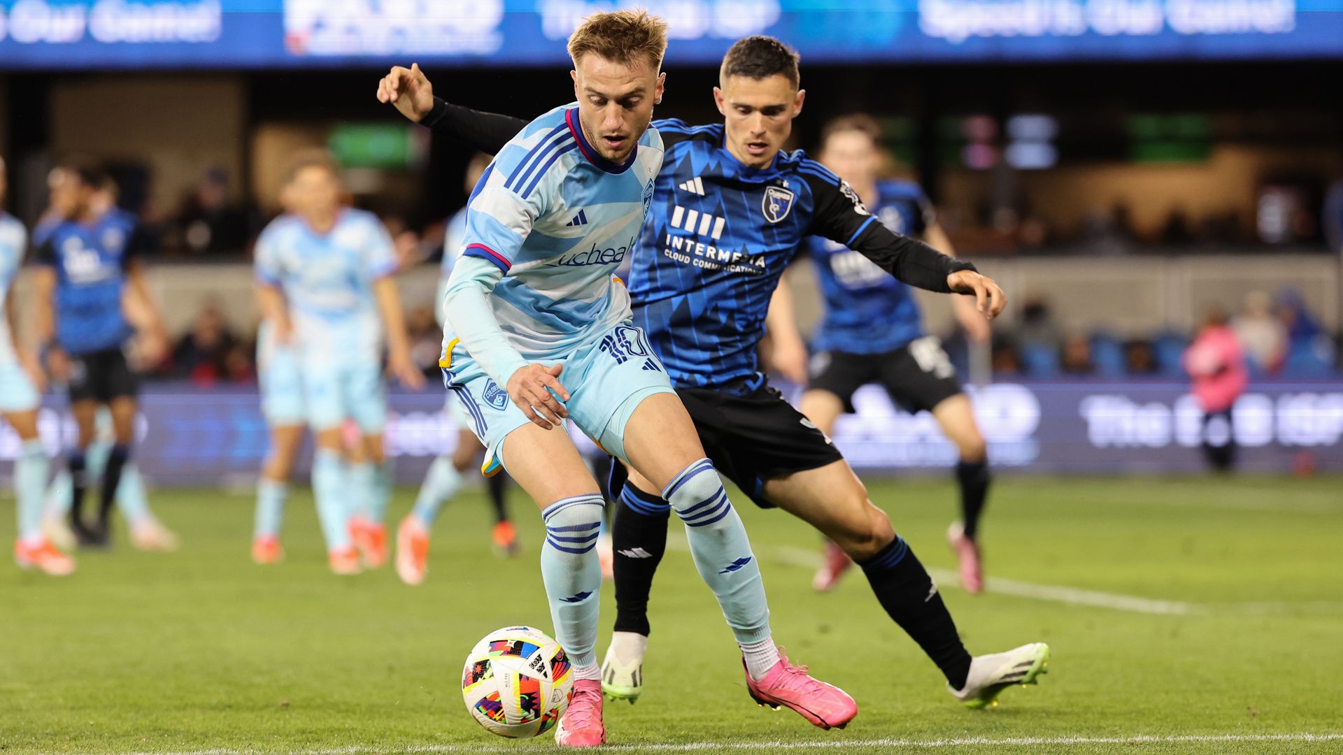 Two soccer players fight for a ball near the feet of a player with a light blue uniform. 