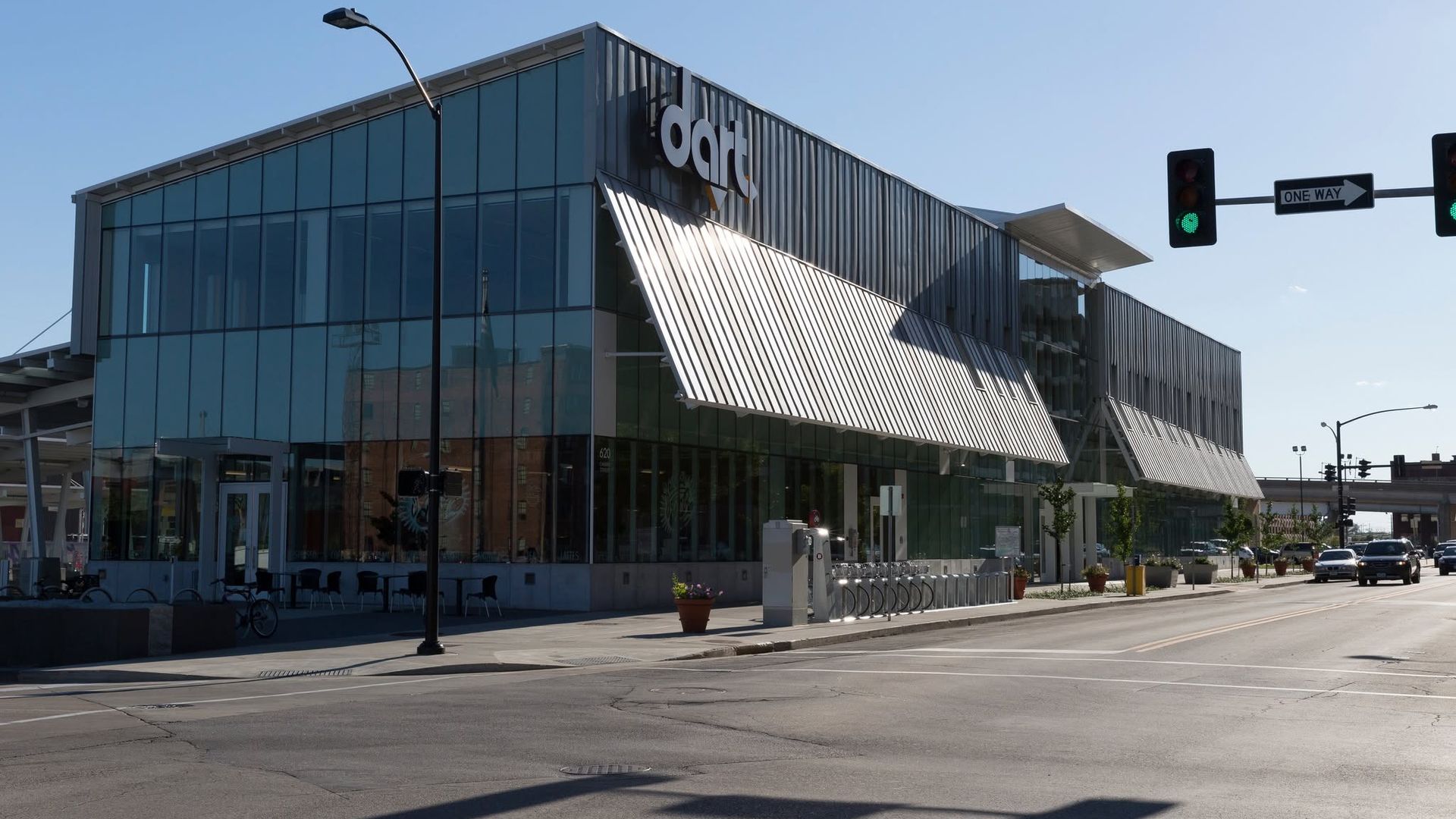 Modern glass building with a silver diagonal sunshade and a bold white "ODOT" sign on the facade, street traffic signals and a one-way arrow sign against a clear blue sky.