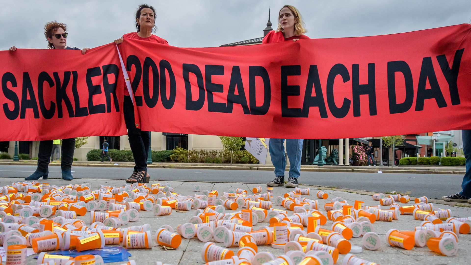 In this image, a banner reading "Sackler" and "200 each day" is held by three people who stand next to a large number of prescription bottles clustered on the ground.