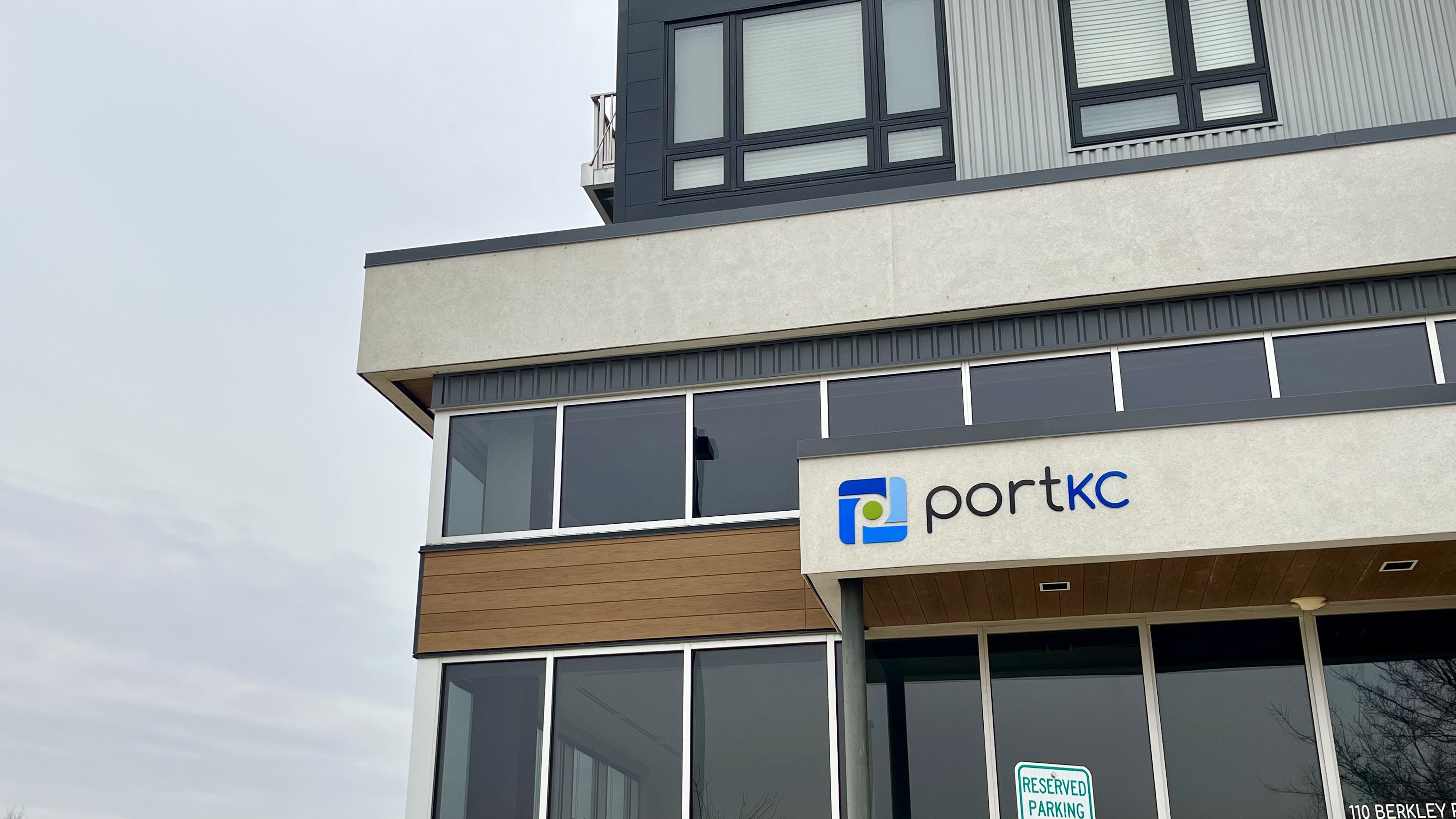 Modern building exterior with gray, wood, and black panels featuring large windows and a sign reading "portkc" with a blue and green logo.