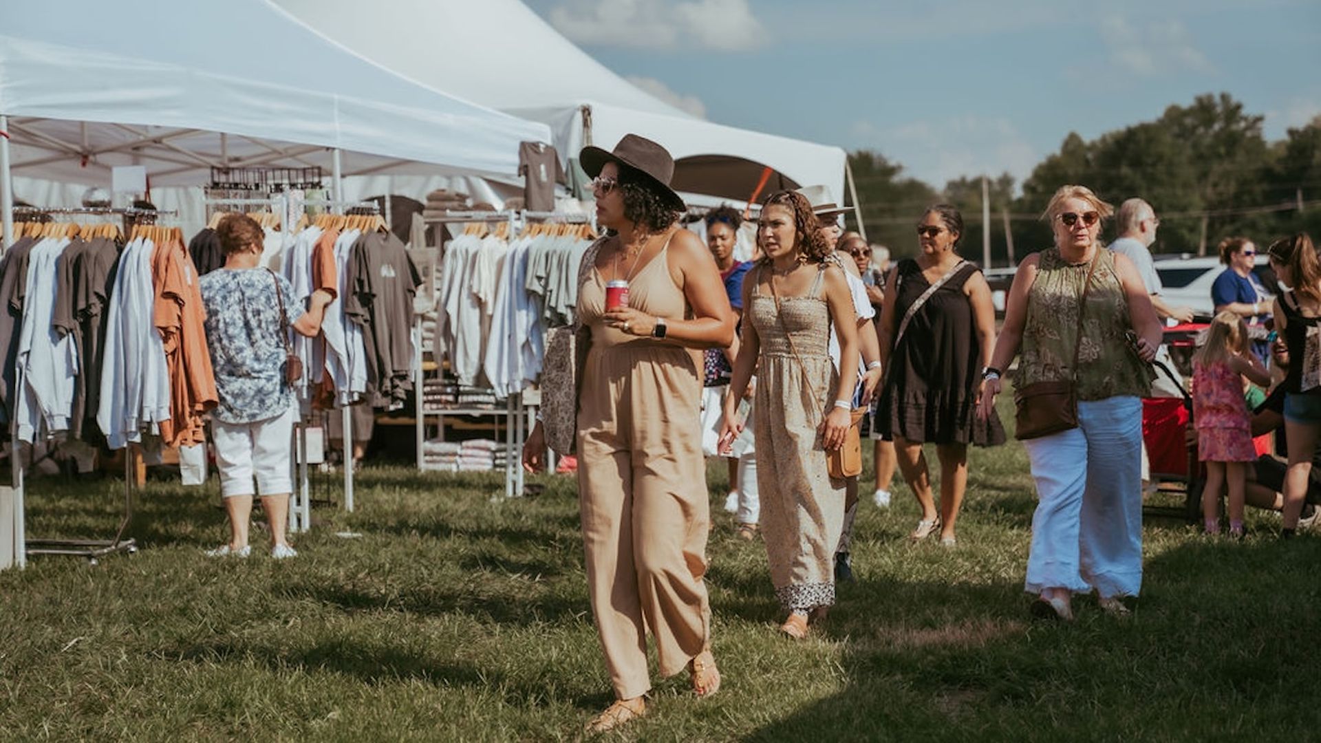 Outdoor clothing market under white canopies; racks of shirts in blues, creams, and browns. Shoppers walk on grass, including a woman in a tan jumpsuit and hat holding a red cup.
