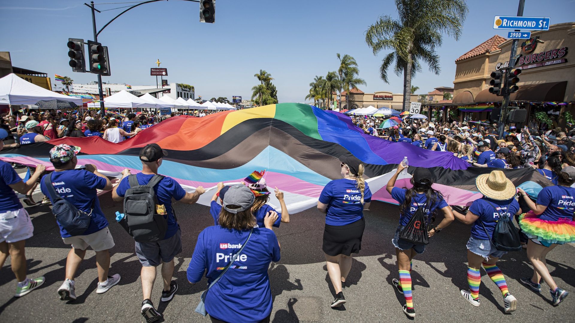A group of people in Pride tee shirts and rainbow accessories march down the street holding a large Progress Pride flag during a parade.