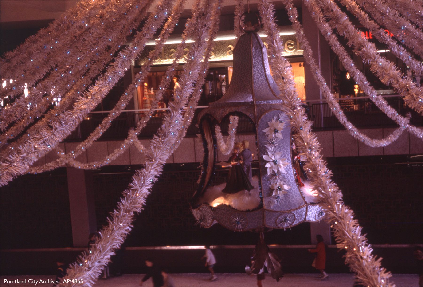 Large decorative bell ornament hanging with white garland strands and lights in a mall, with people walking below and store windows in the background.