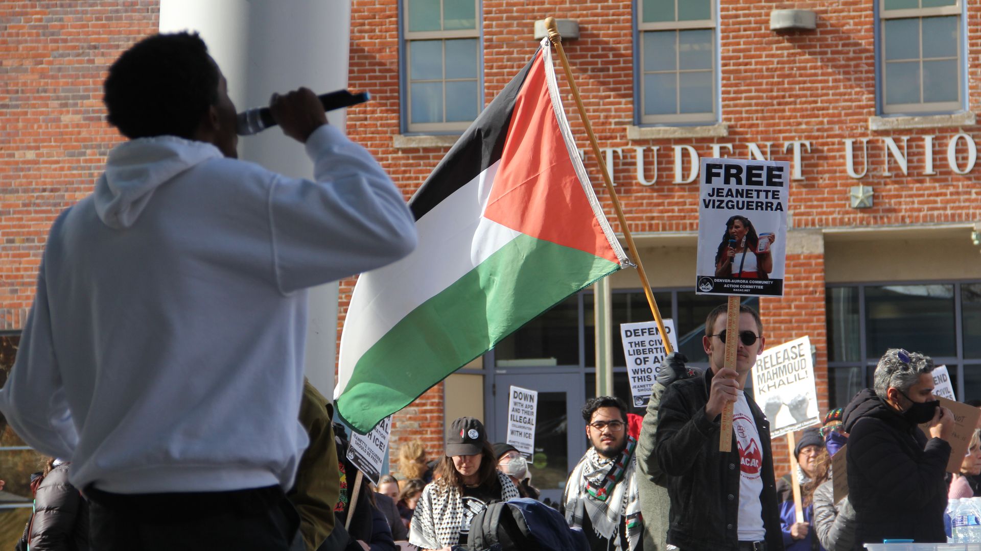 A man holds up a large Palestinian flag amid a crowd outside a large brick building. 