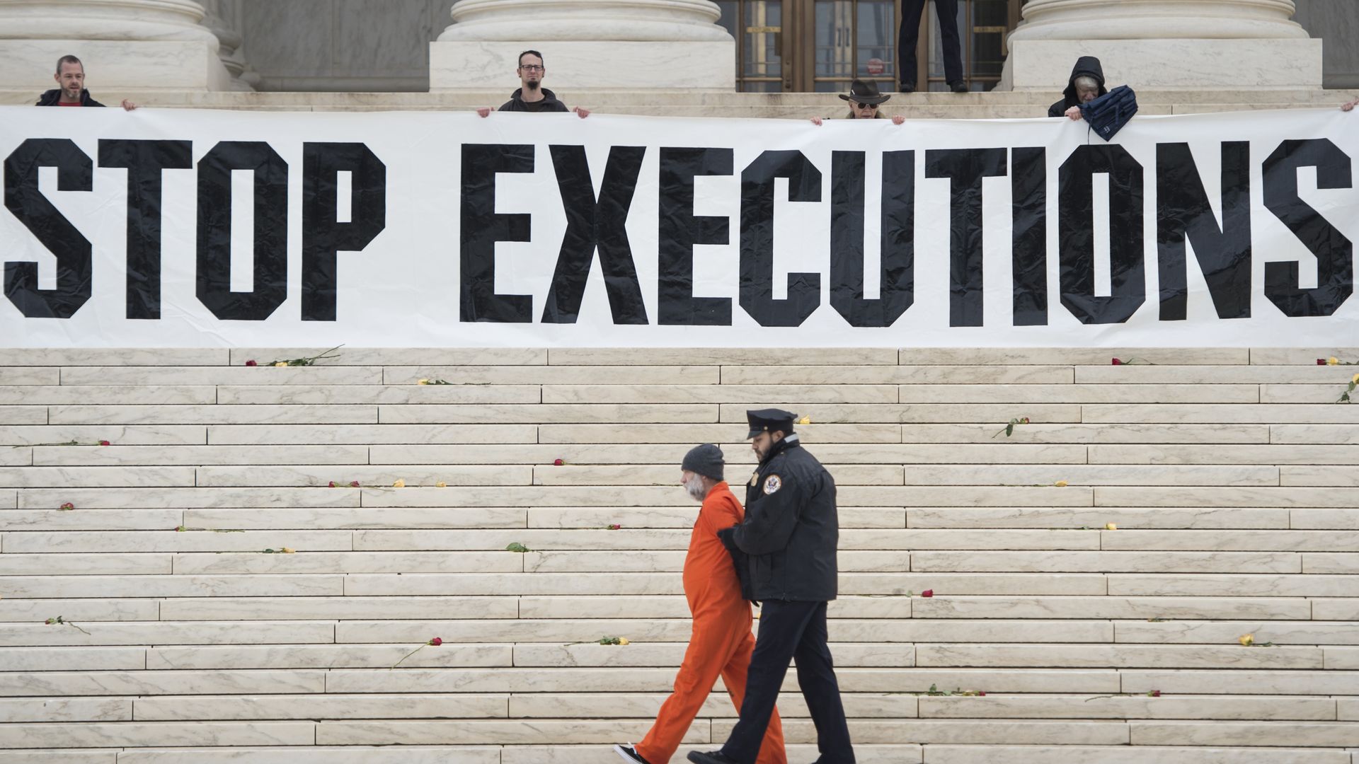 man in orange jumpsuit being escorted by police officer sign that says "stop executions" is being held in the background