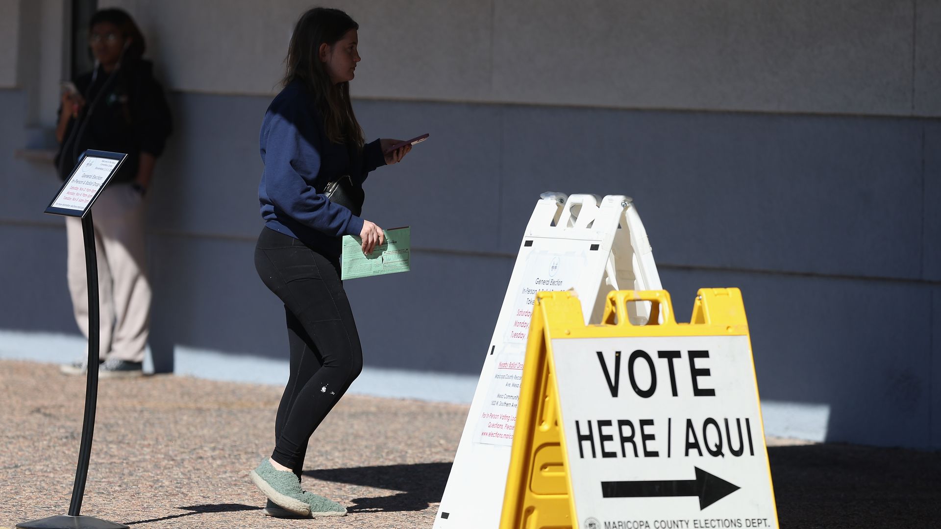 A young woman holding a green paper and phone approaches signs directing voters, including a yellow one that reads "VOTE HERE / AQUI" with an arrow, outside a building.
