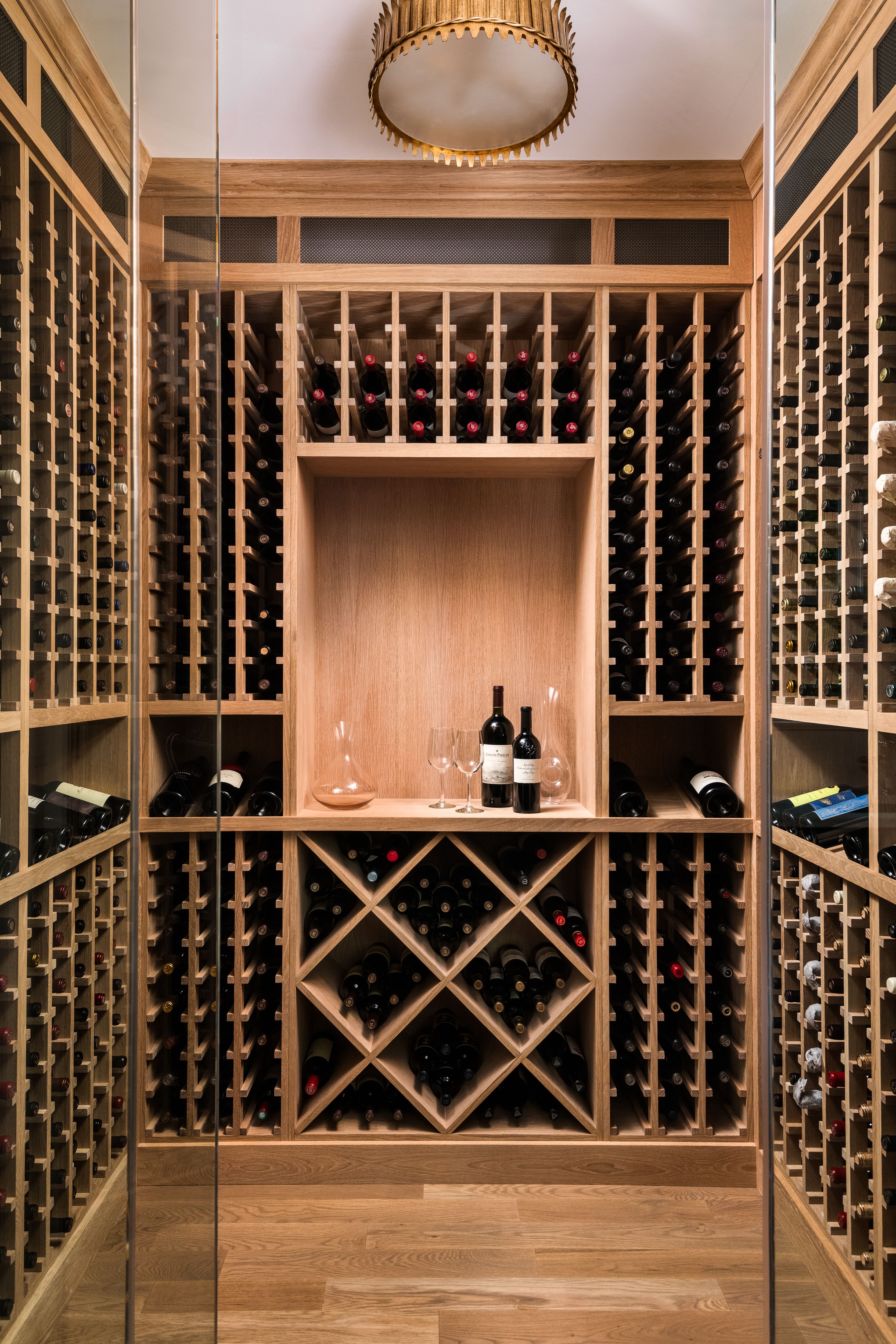 Wooden wine cellar with racks filled with many wine bottles. Two bottles, two glasses, and a decanter displayed in a central cubby. Warm lighting and wood floor.
