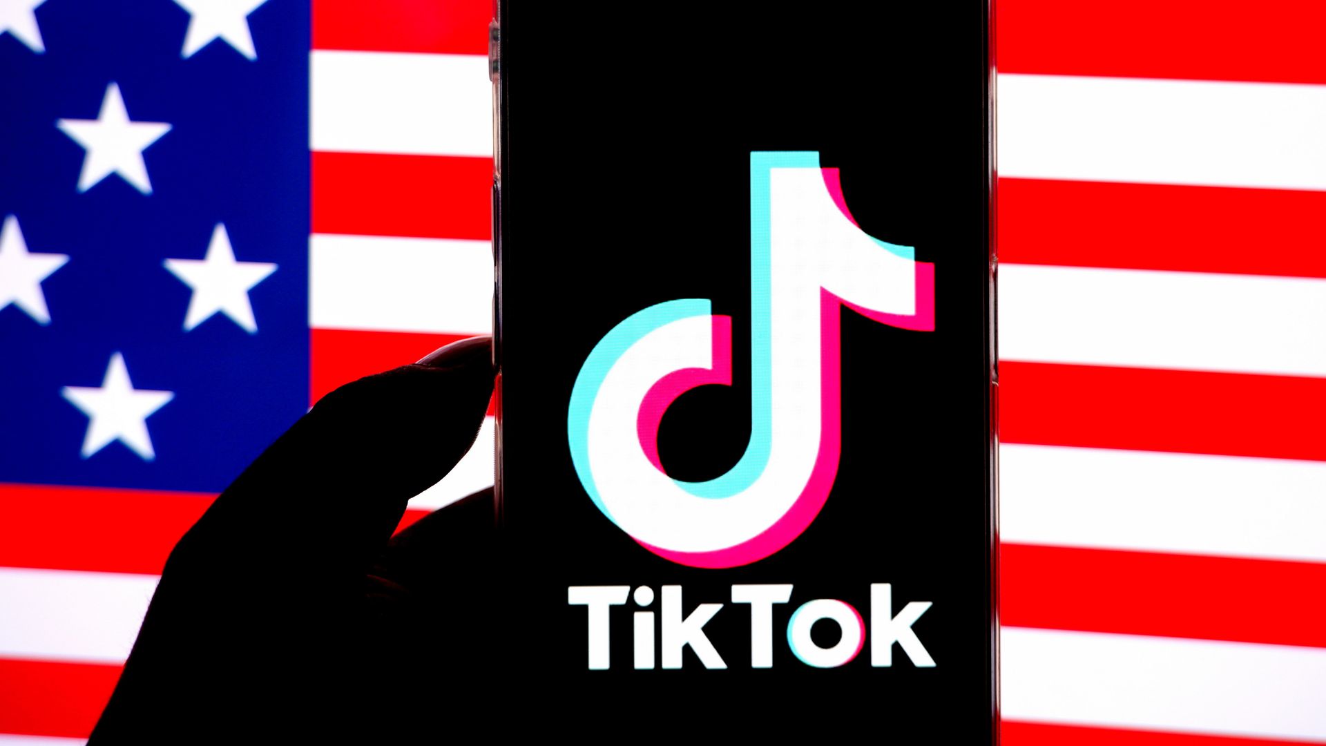 A silhouetted hand holds a phone with the white, pink and blue TikTok logo in front of an American flag.