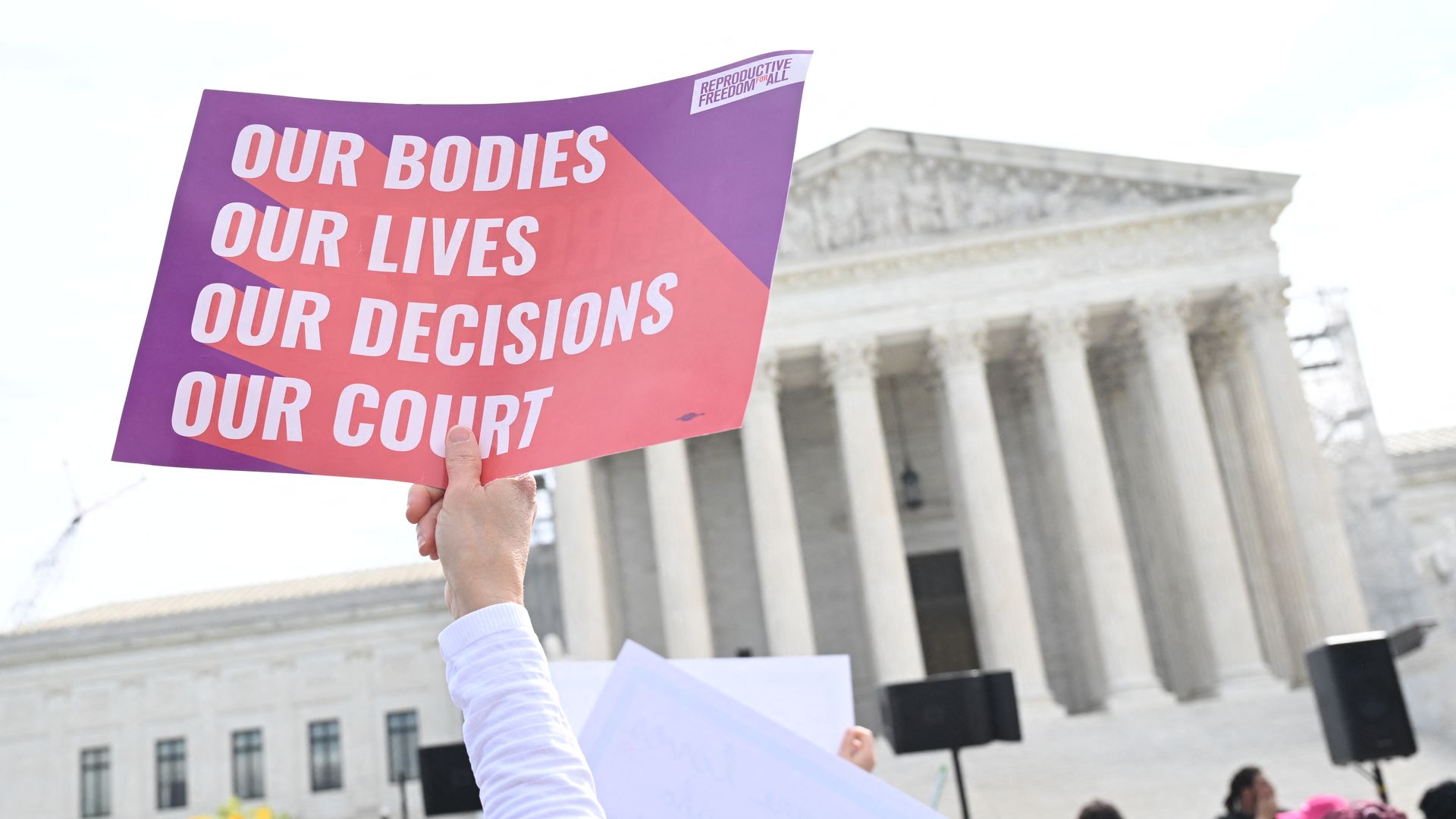 A person holds a sign that reads "our bodies, our lives, our decisions, our court" in front of the Supreme Court, a white building with columns in front.