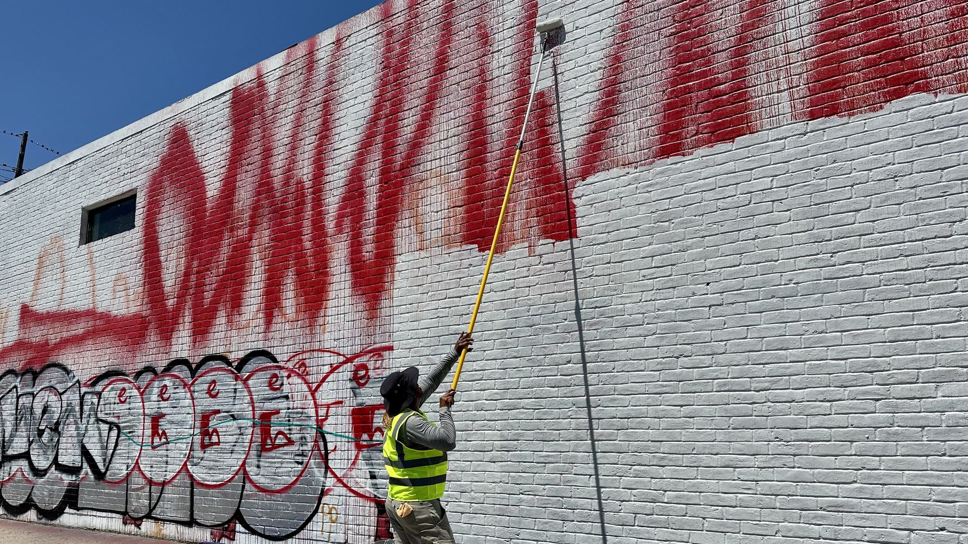 Photo shows a man painting over graffiti on a white wall
