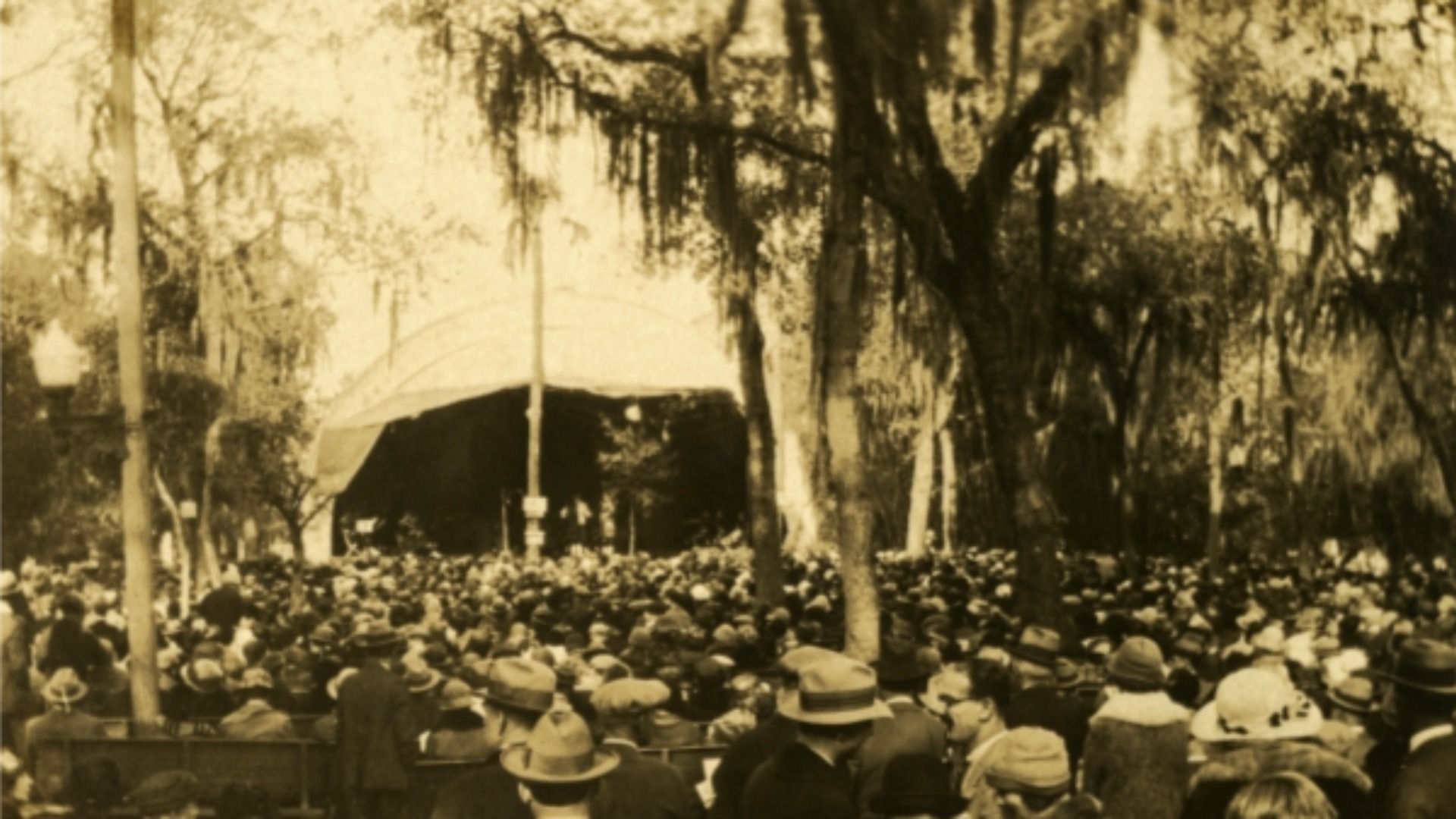 Sepia-toned outdoor scene showing a large crowd seated in a park, facing a raised stage under a white canopy tent, surrounded by tall trees and dappled sunlight.