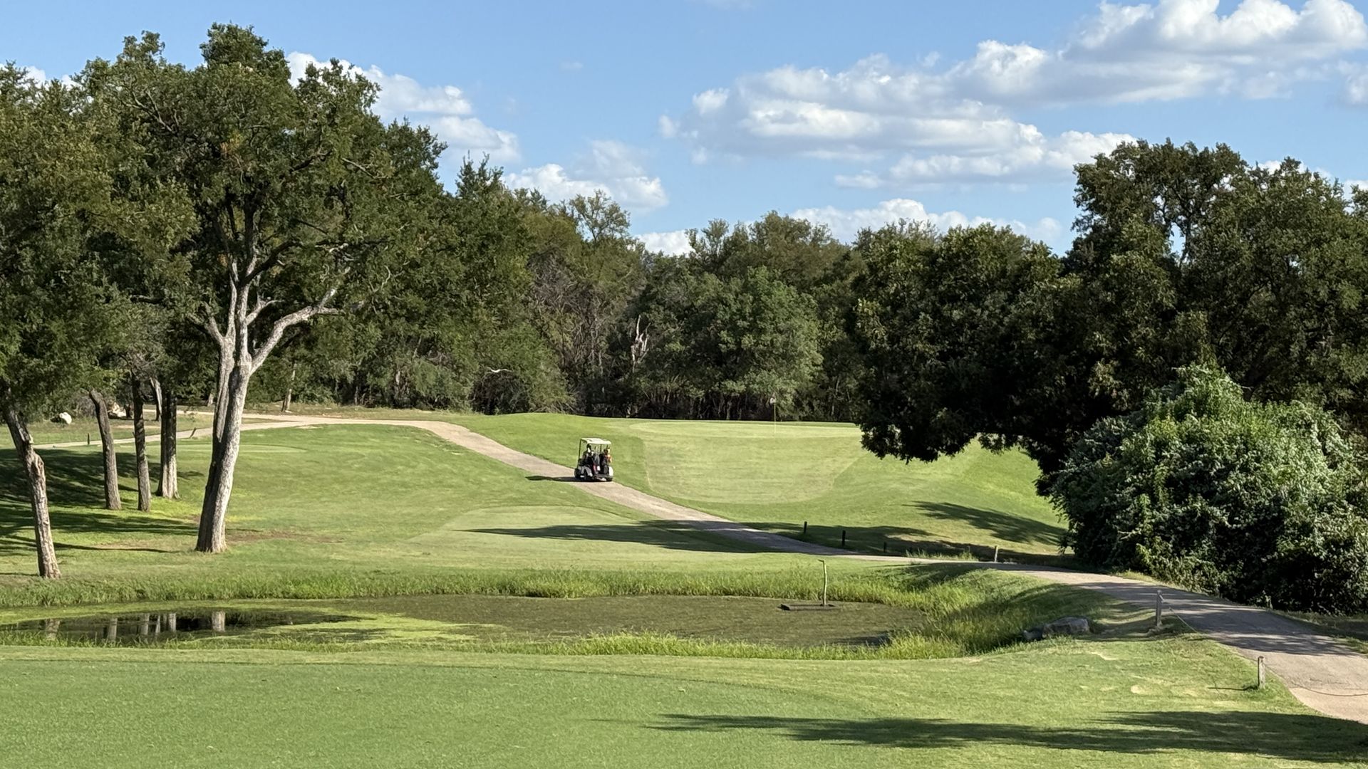 Sunny golf course with a winding cart path, dense trees on both sides, and a small pond in the foreground. A two-person golf cart sits mid-path under a blue sky with fluffy clouds.