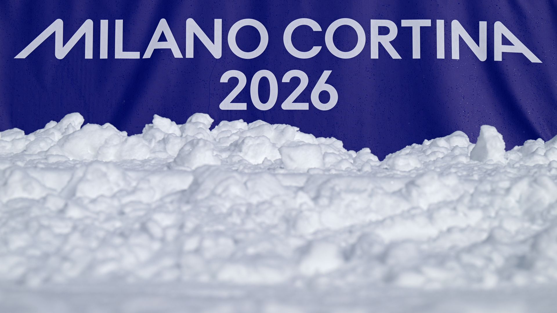 White snow in the foreground with a blue banner behind it displaying "MILANO CORTINA 2026" in large white letters.
