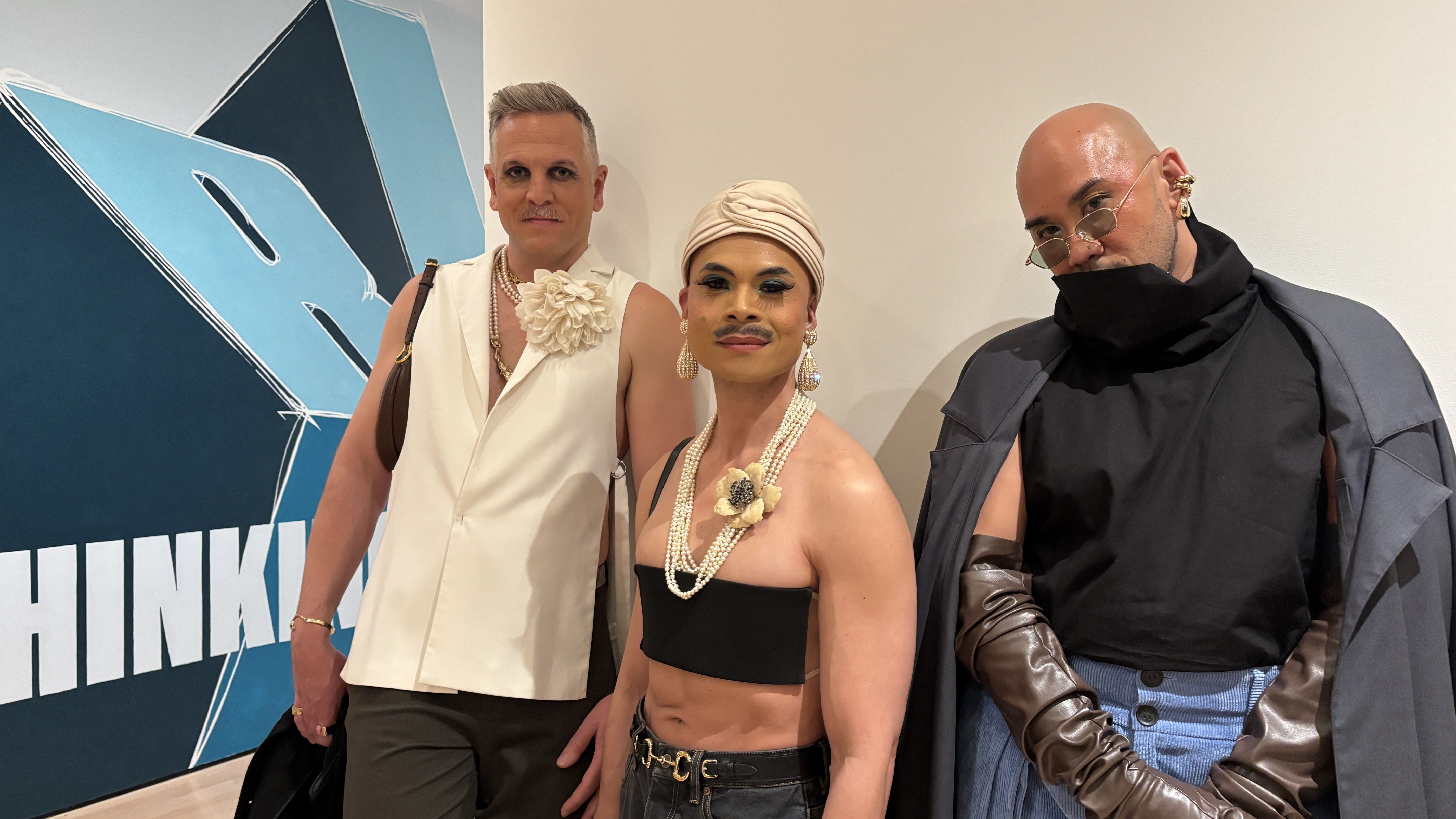 Three stylish people pose indoors before a blue geometric mural: left man in white sleeveless with a flower and pearls; center person in a beige turban with bold makeup; right bald person in a dark cape and glasses.
