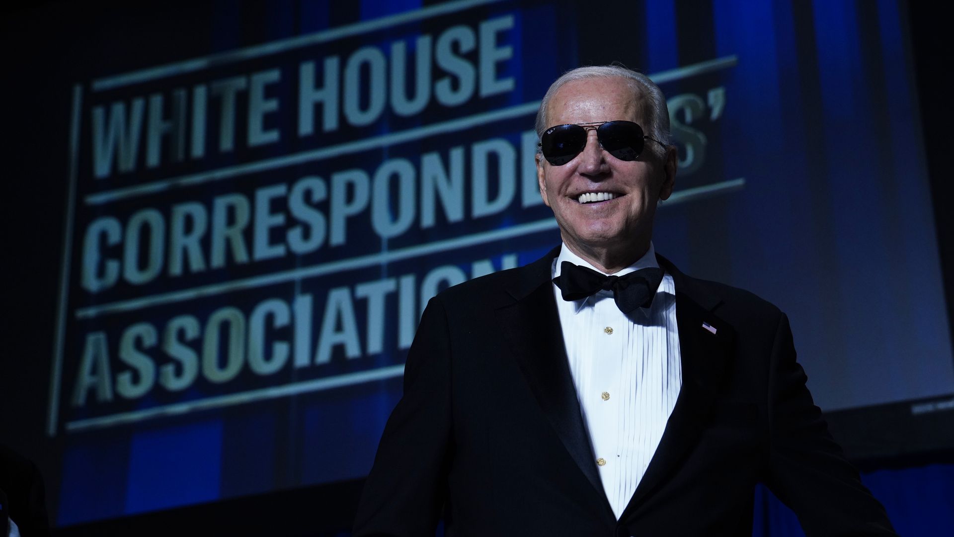 President Biden wearing sunglasses while standing in front of a White House Correspondents' Association sign.