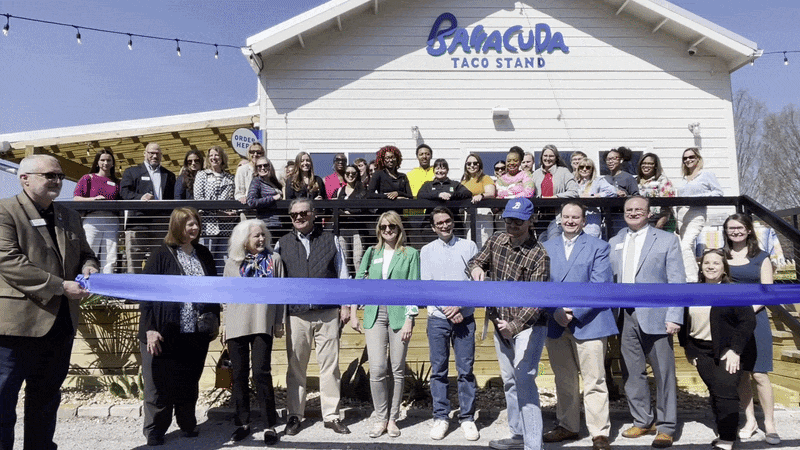 A diverse group stands in front of a white building with a blue sign reading Barracuda Taco Stand, a purple ribbon stretched across for a ribbon-cutting, under string lights.