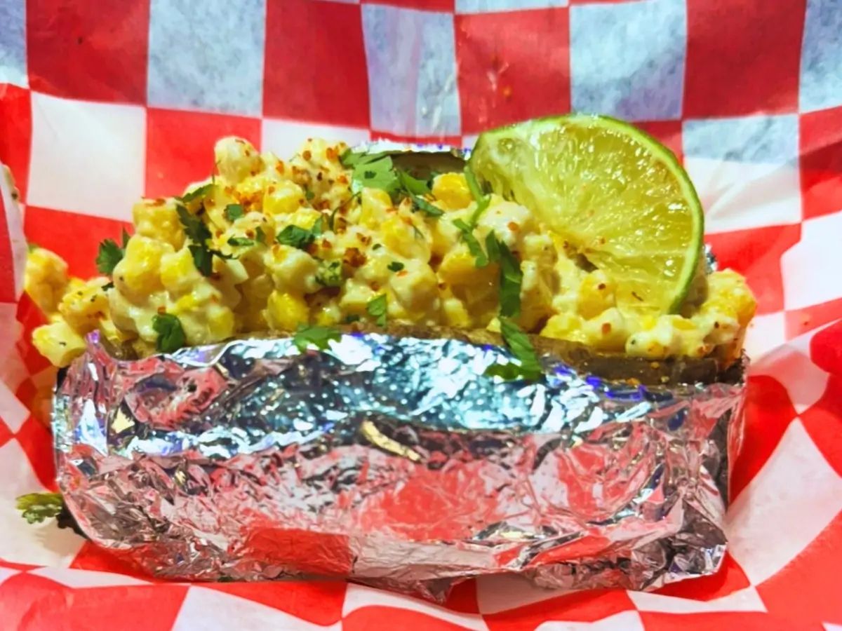 A pile of corn with cilantro and a lime on top tucked in a baked potato wrapped in tin foil and sitting on a white- and red-checkered piece of butcher paper.