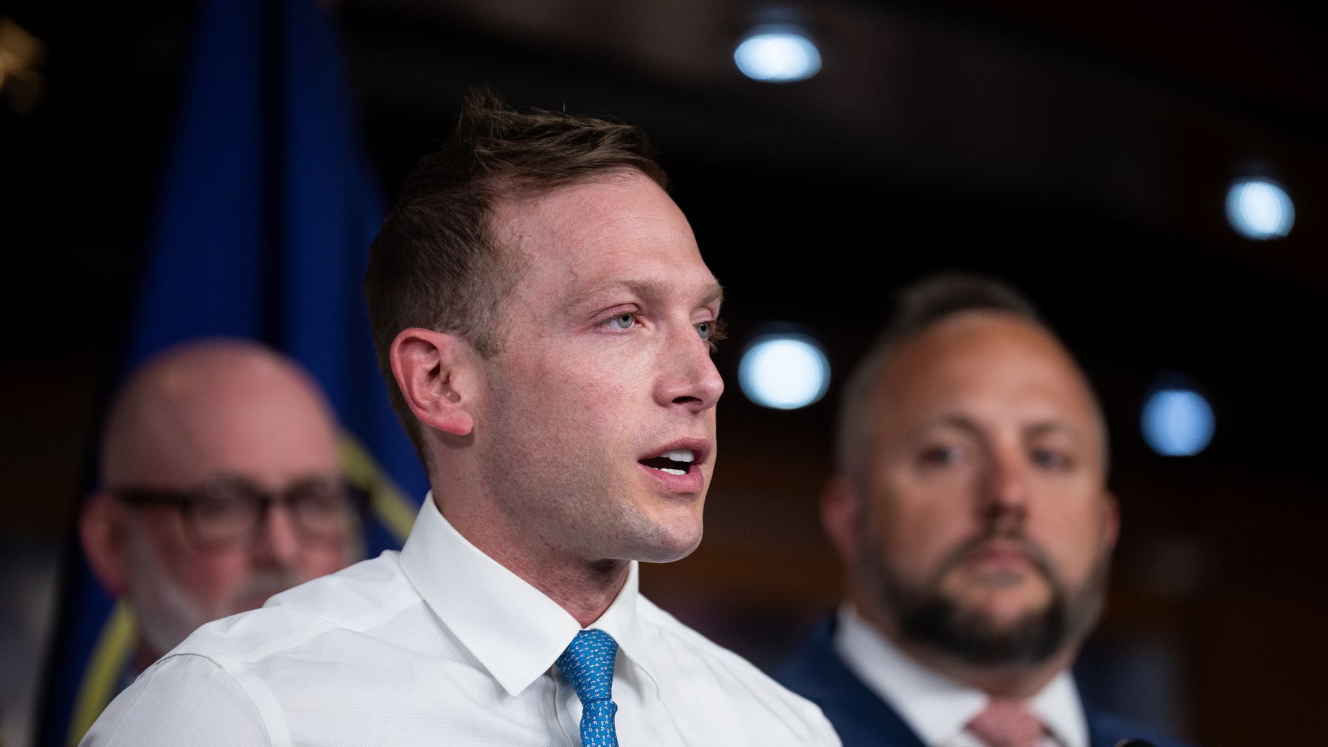 Close-up of a man in a white shirt and blue tie (Max Miller) speaking at a press conference, microphones in front, blurred colleagues behind, and a blue flag visible on the left.