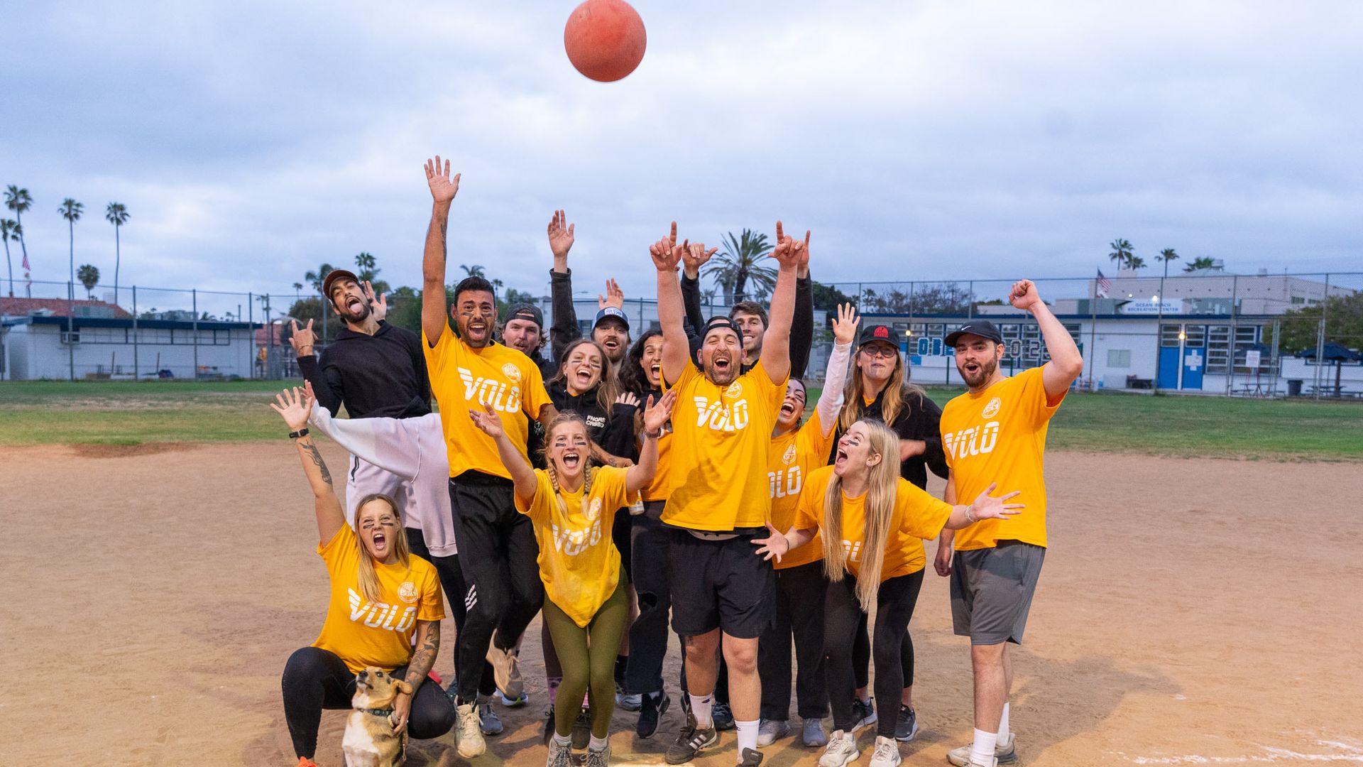 A kickball team in yellow jerseys celebrates on the infield dirt, while throwing a ball in the air