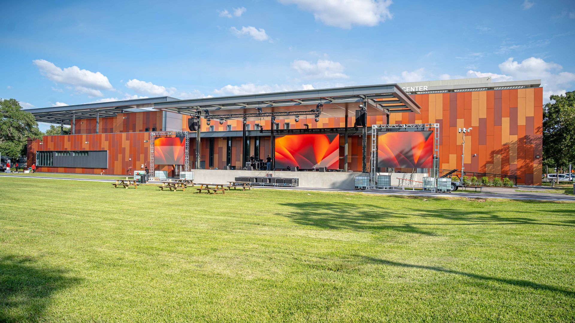 Outdoor concert stage in front of a modern orange-panel building under a blue sky, with large video screens, metal trusses, speakers, and a grassy field with picnic tables.