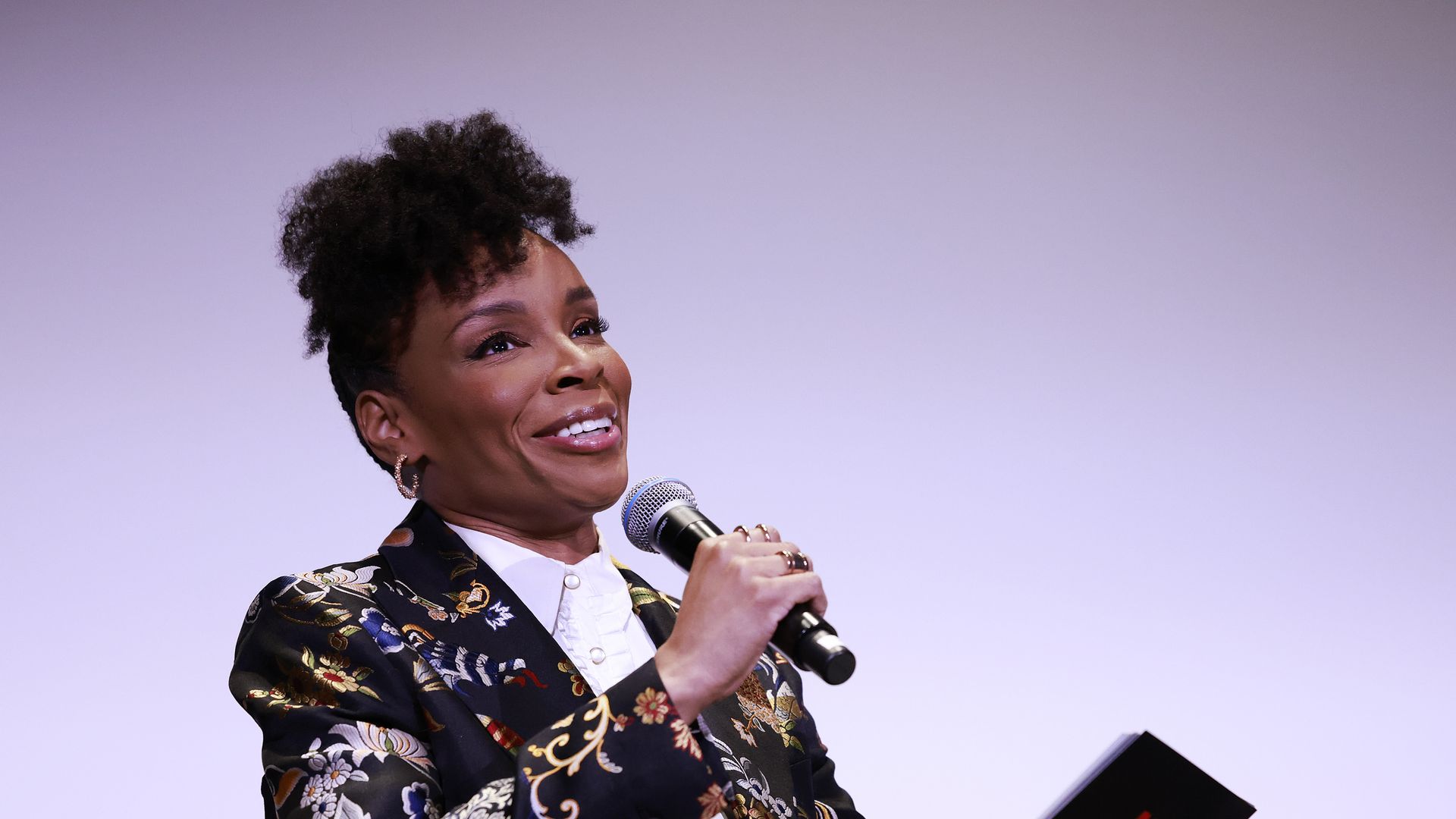 A photo of the comedian Amber Ruffin sitting in a chair and holding a microphone.