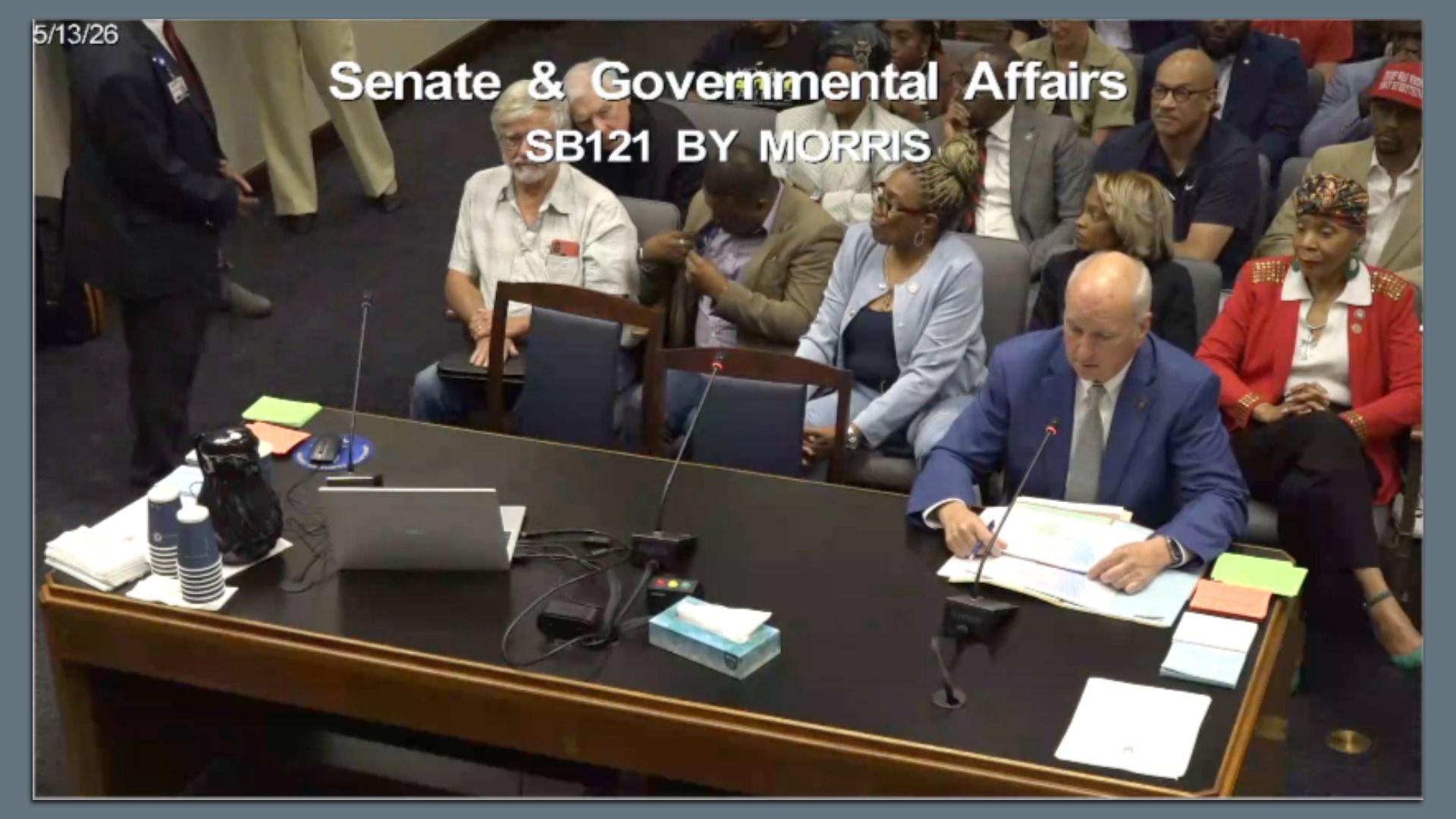 Formal hearing room; a man in a blue suit speaks into a microphone at a desk with papers while an audience watches. Overlay reads "Senate & Governmental Affairs SB121 BY MORRIS".