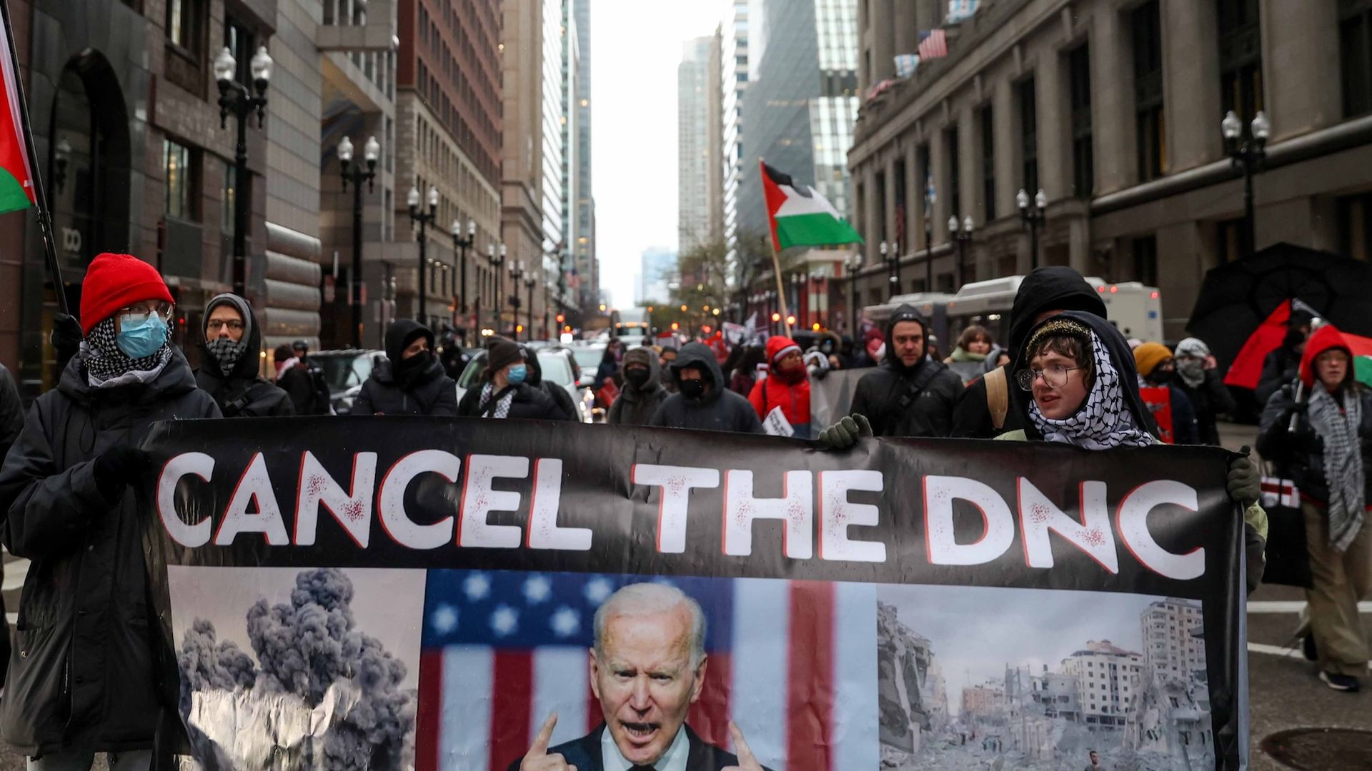 Protesters carry a banner that reads "CANCEL THE DNC" with photos of bomb smoke, President Biden in front of a flag, and a Gaza. They are leading other protesters on a Chicago street.
