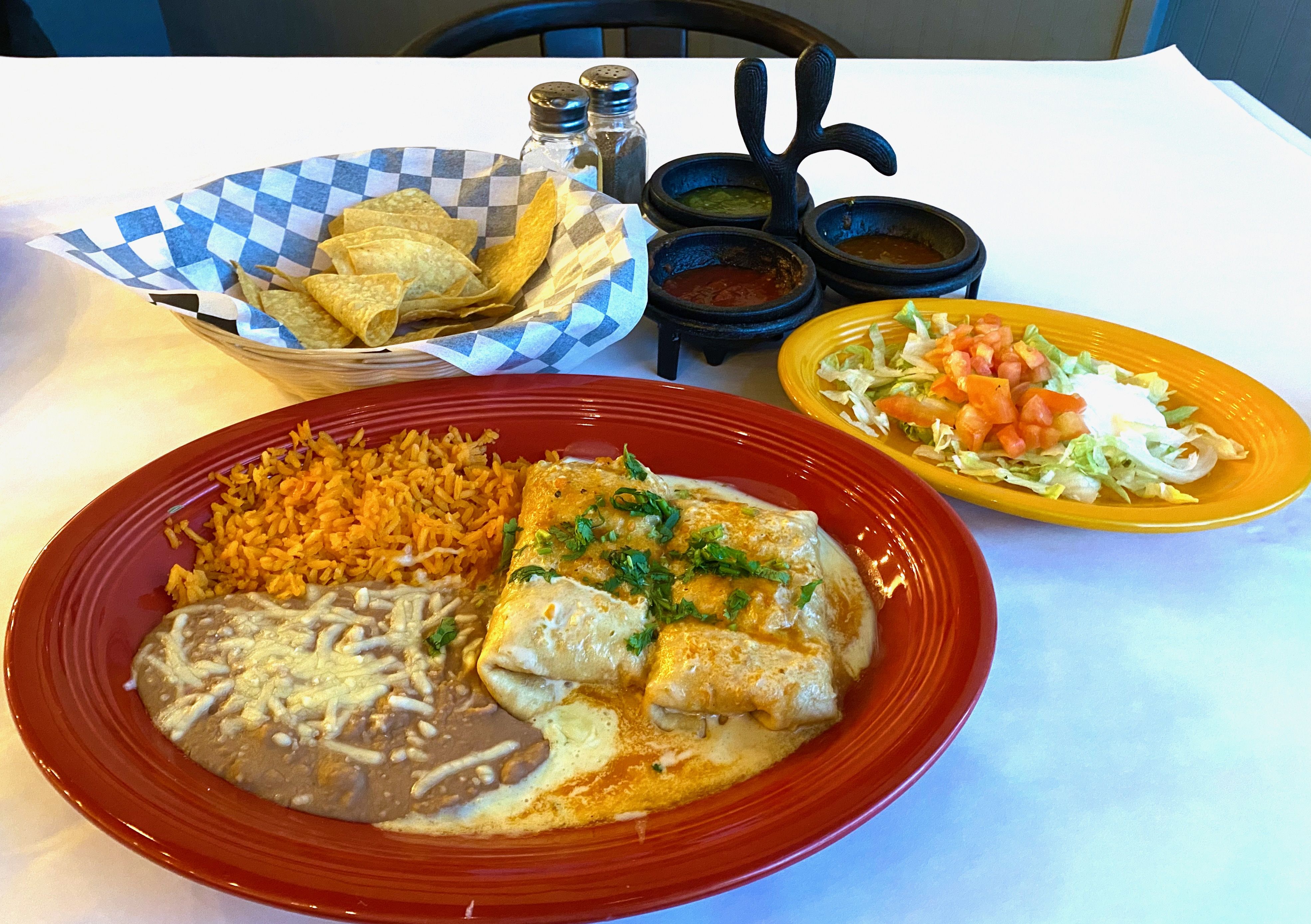 A plate with chimichangas, rice and peans, a plate with fajita salad, and a bowl with chips next to three salsas in a holder