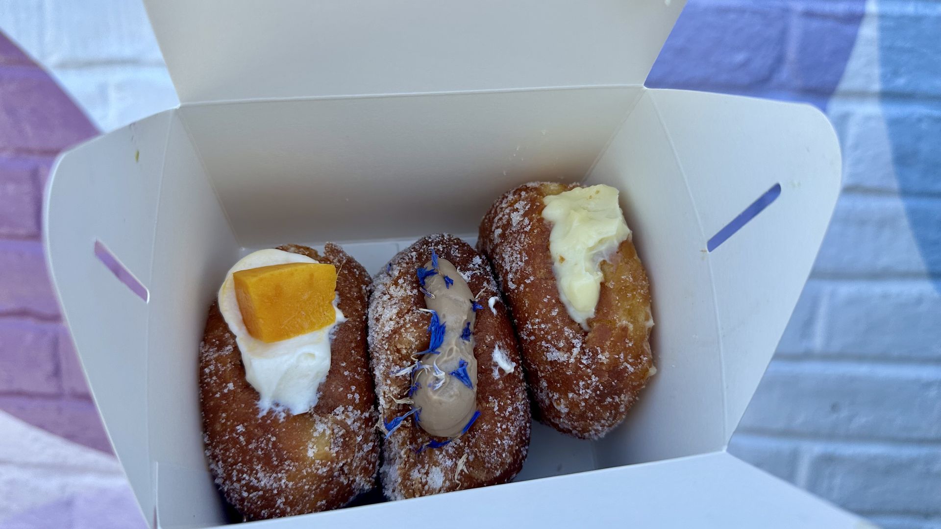 White takeout box holds several sugar-dusted doughnuts topped with cream, a mango slice, and blue edible flowers, set on a pastel background.