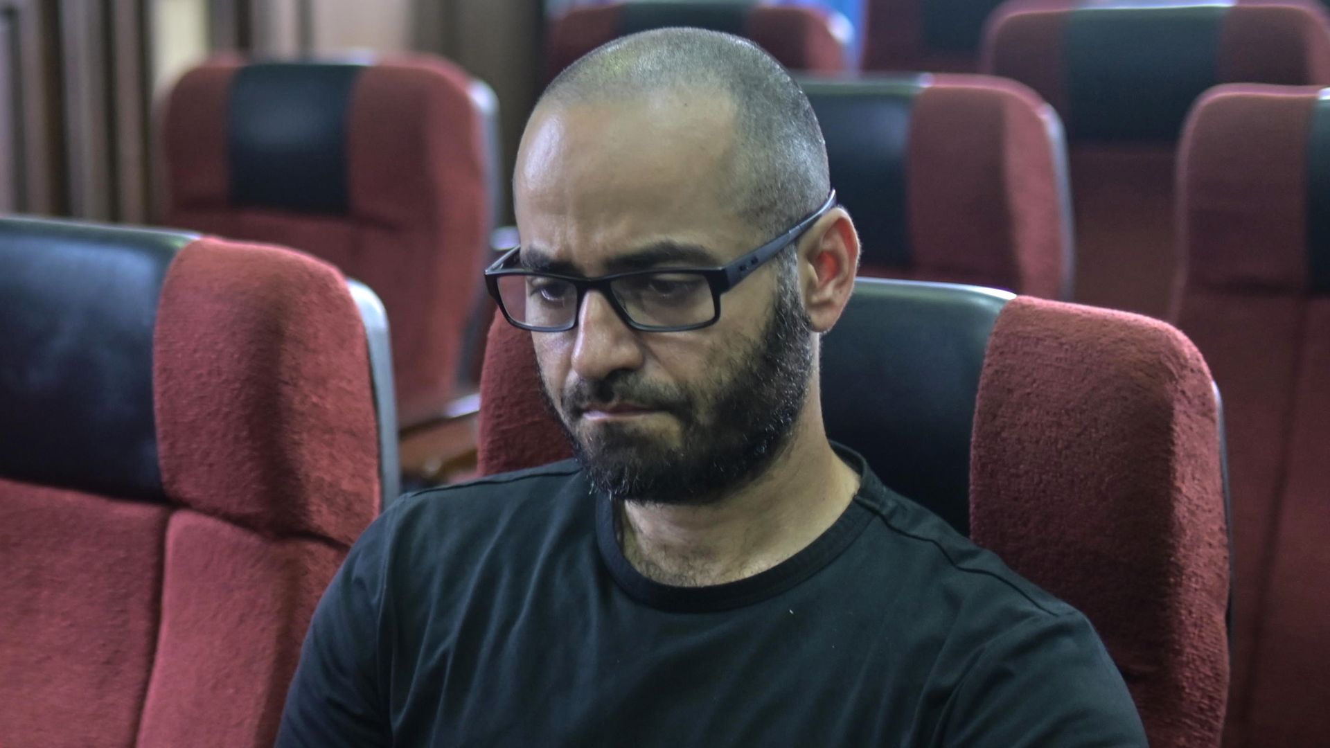 A man in a black tshirt, light beared and glasses sitting in a sort of gallery, with sunlight coming in from a window behind him. 