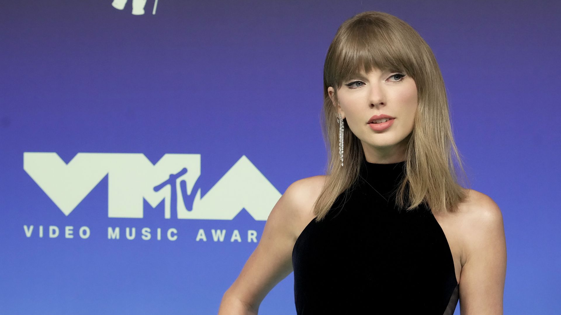 A woman with long light brown hair in a black sleeveless dress stands against a blue to purple gradient backdrop with a large white MTV logo; she wears a long sparkling earring.